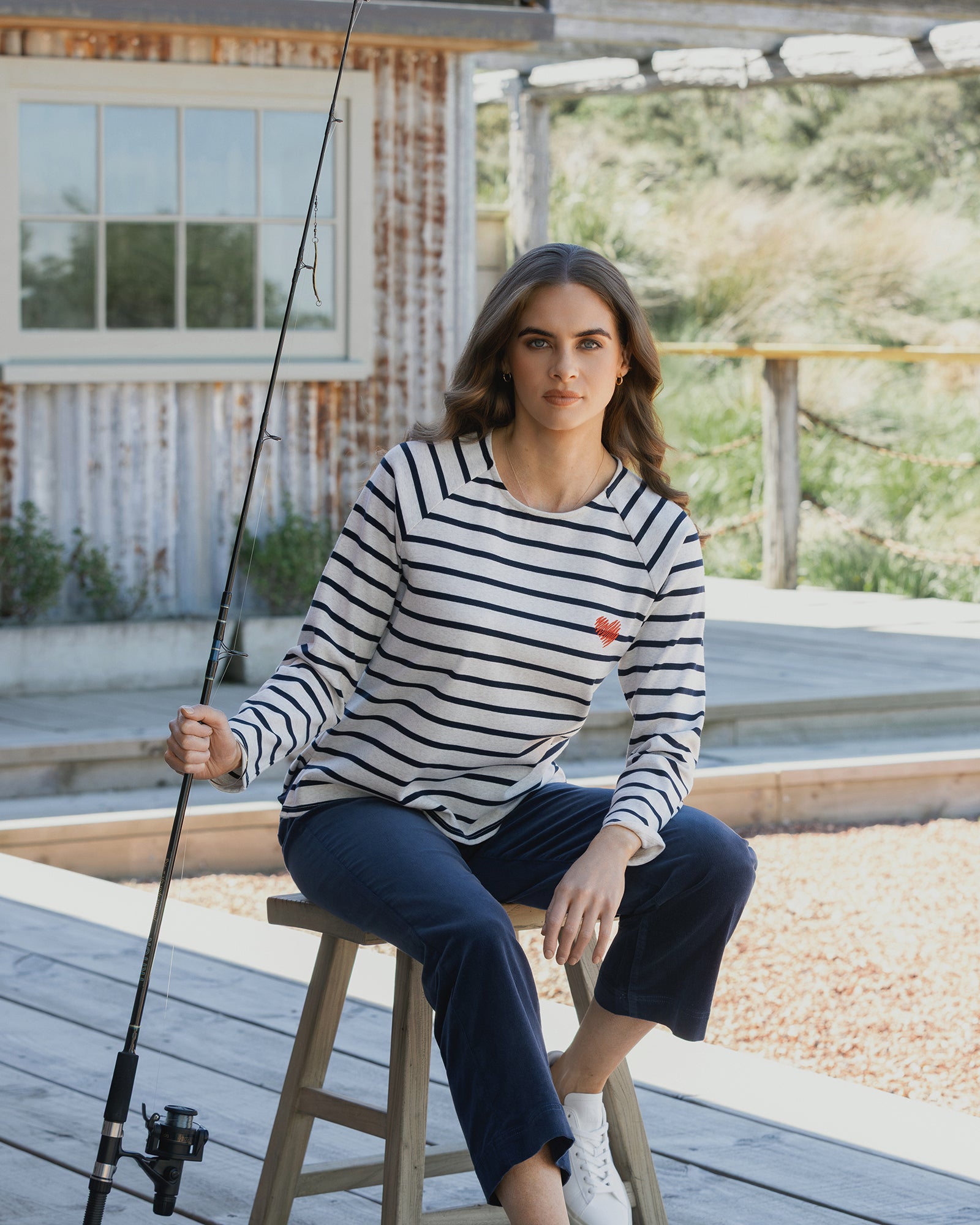A woman wearing the Vassalli Lover Top in Oat/Ink and dark pants sits on a wooden stool holding a fishing rod, with a rustic building and greenery in the background.