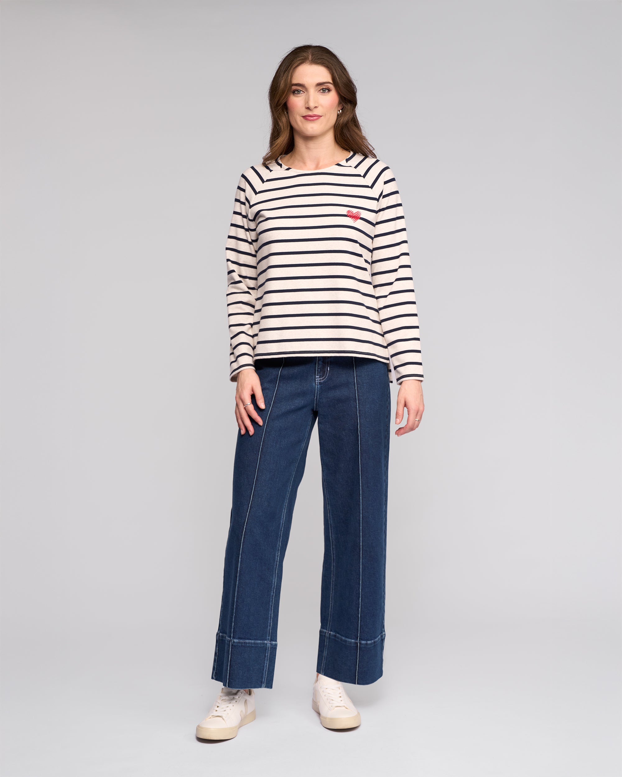 A woman faces forward against a light gray background, wearing the Vassalli Lover Top in Oat/Ink— a cream and black striped long-sleeve cotton jersey with a small red heart patch—paired with wide-leg blue jeans and white sneakers.