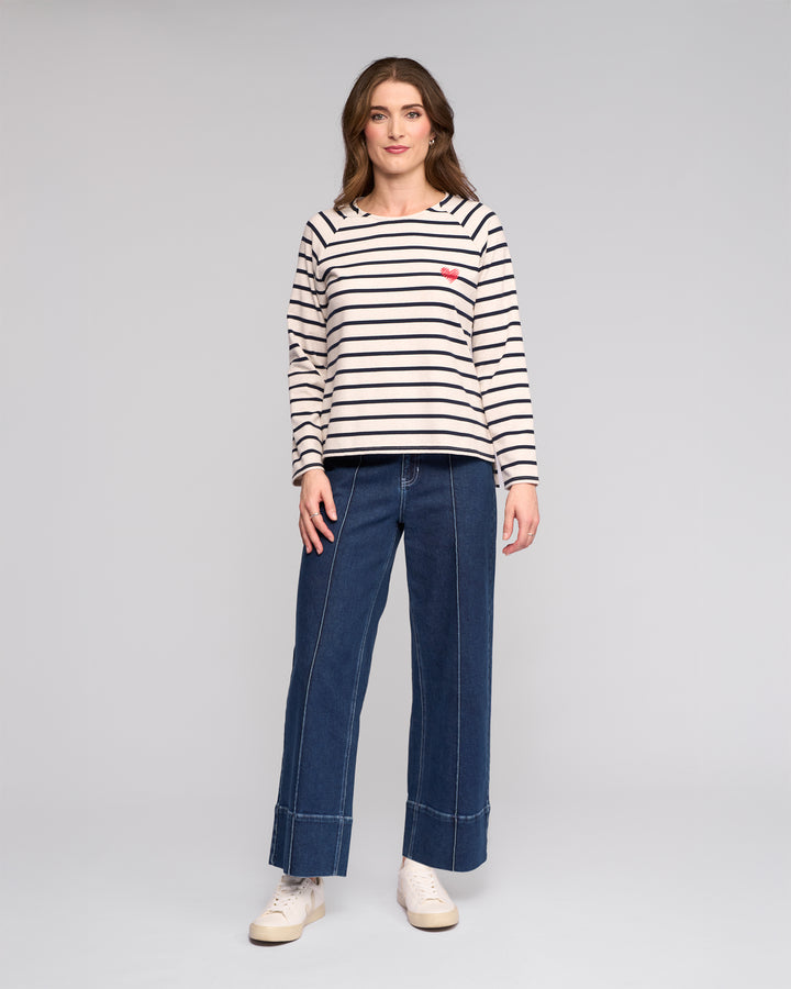 A woman faces forward against a light gray background, wearing the Vassalli Lover Top in Oat/Ink— a cream and black striped long-sleeve cotton jersey with a small red heart patch—paired with wide-leg blue jeans and white sneakers.