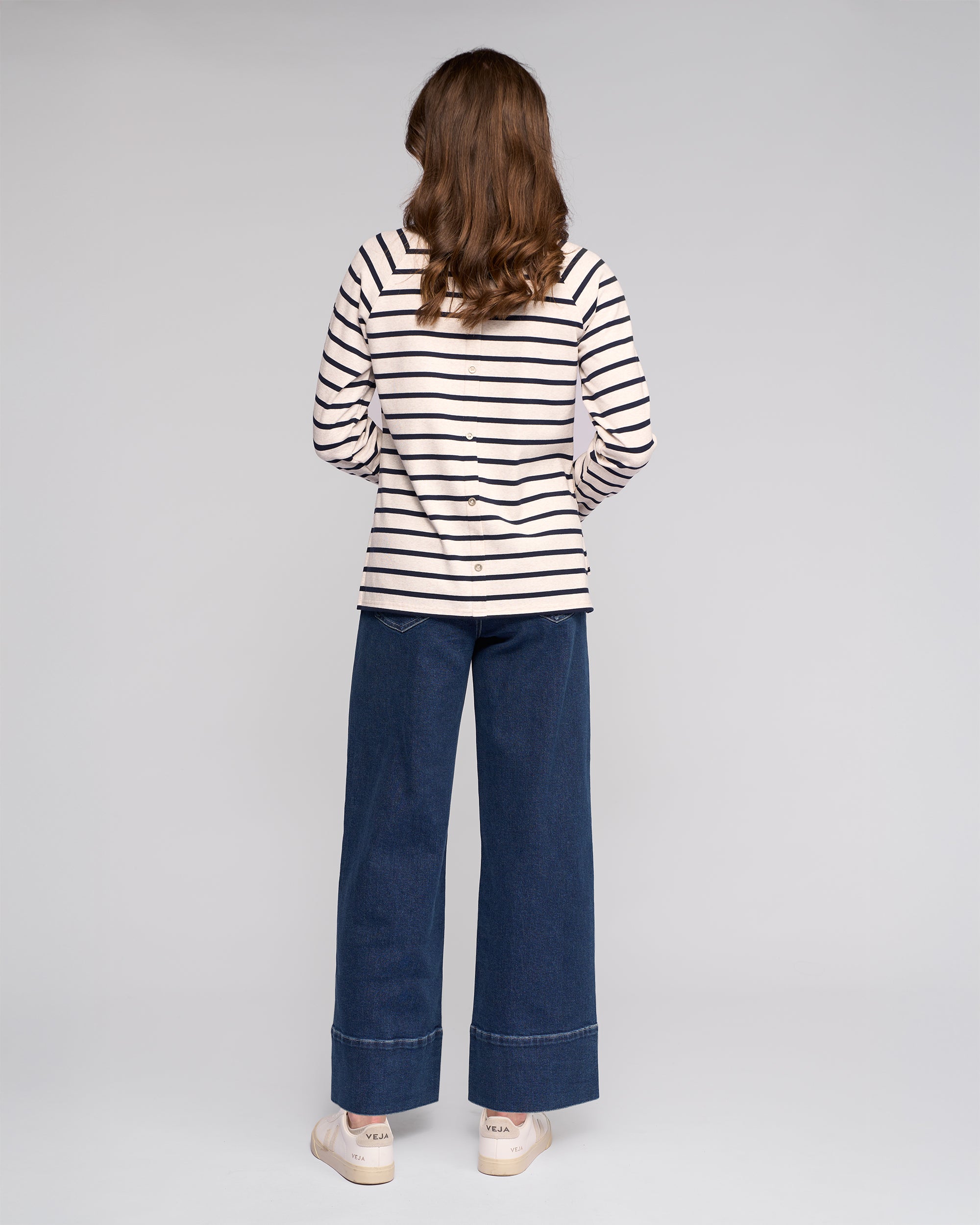A person with long brown hair wears the Vassalli Lover Top in Oat/Ink—an oat and ink striped cotton jersey long-sleeve—paired with wide-leg blue jeans and white sneakers, standing against a plain light gray background.