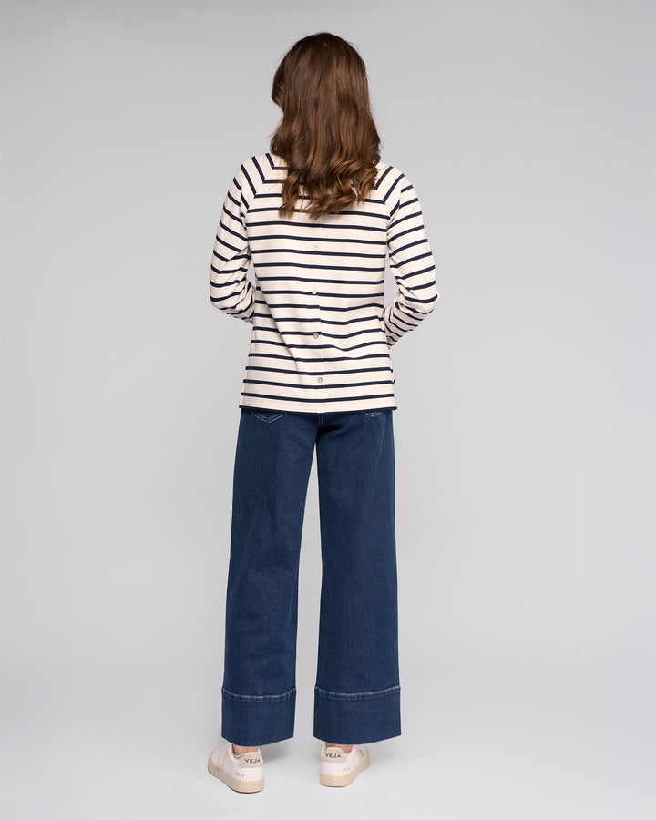 A person with long brown hair wears the Vassalli Lover Top in Oat/Ink—an oat and ink striped cotton jersey long-sleeve—paired with wide-leg blue jeans and white sneakers, standing against a plain light gray background.