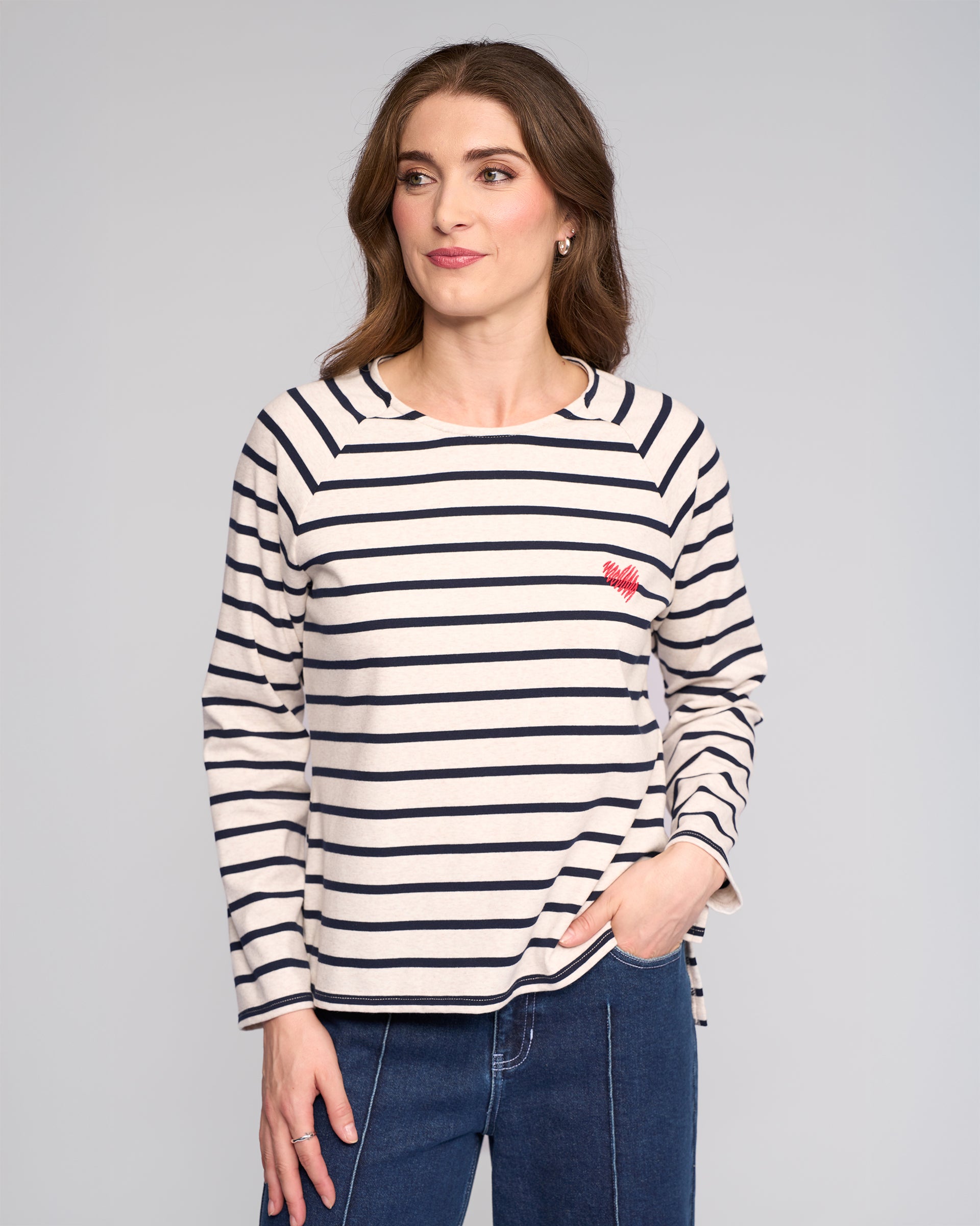 A woman with wavy brown hair wears the Vassalli Lover Top in Oat/Ink, featuring cream and navy stripes with a red heart on the chest, paired with blue jeans. She stands against a light background, looking right and smiling slightly.
