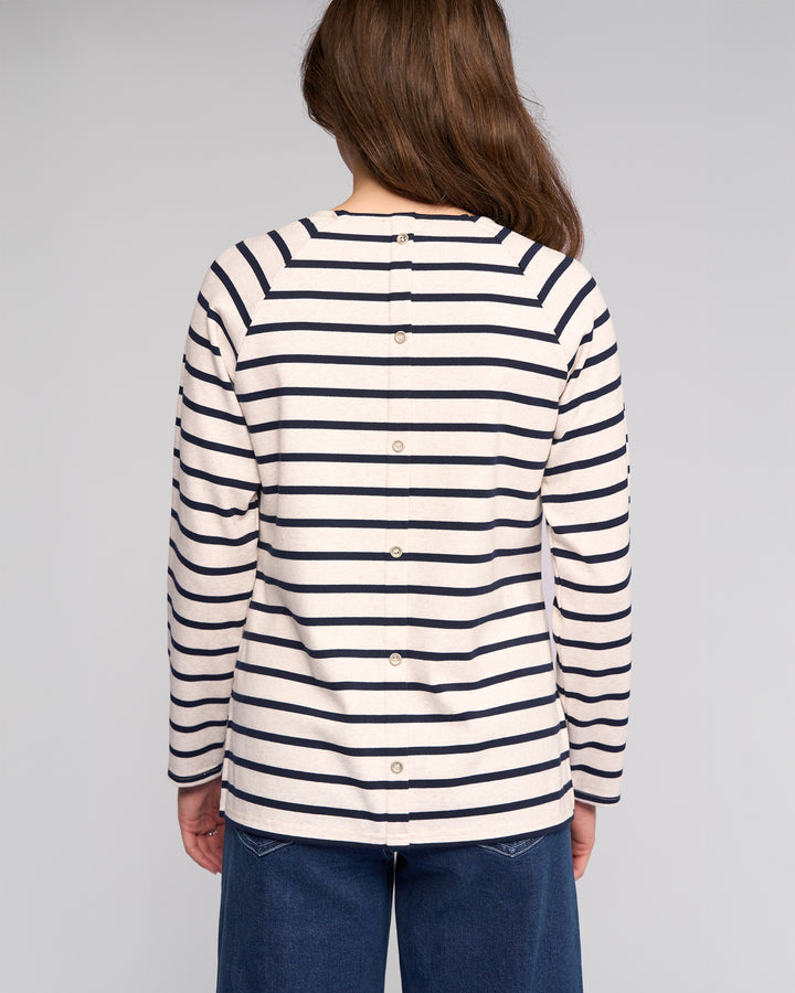 A person with long brown hair faces away, wearing the Vassalli Lover Top - Oat/Ink, a cream cotton jersey long-sleeve with black stripes and back buttons, paired with blue jeans. The background is plain and light-colored.