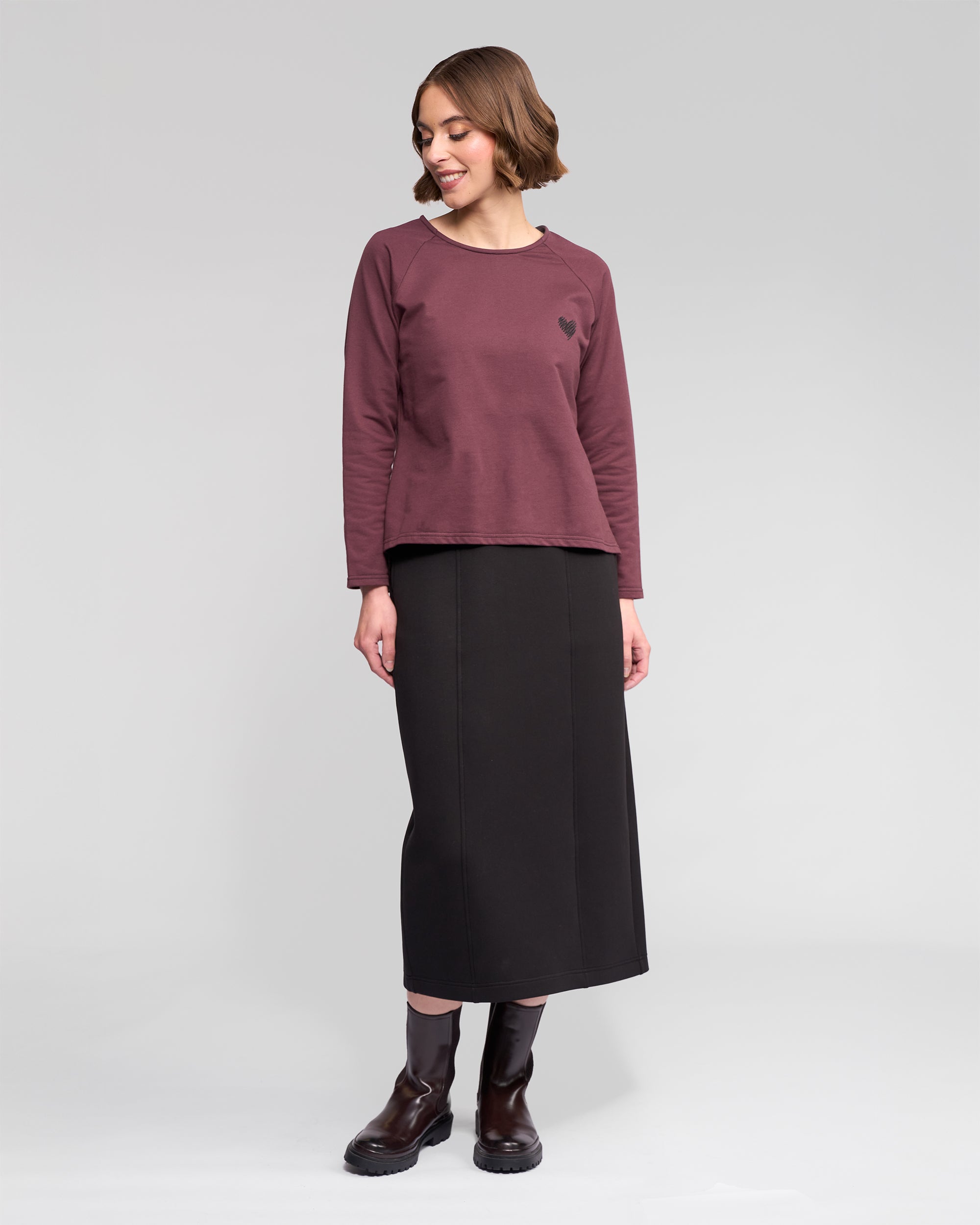 A woman stands in a studio wearing the Vassalli Lover Top - Port, a maroon relaxed long-sleeve with a small heart graphic, paired with a long black skirt and black boots. She looks down, smiling, her short brown hair styled in waves.
