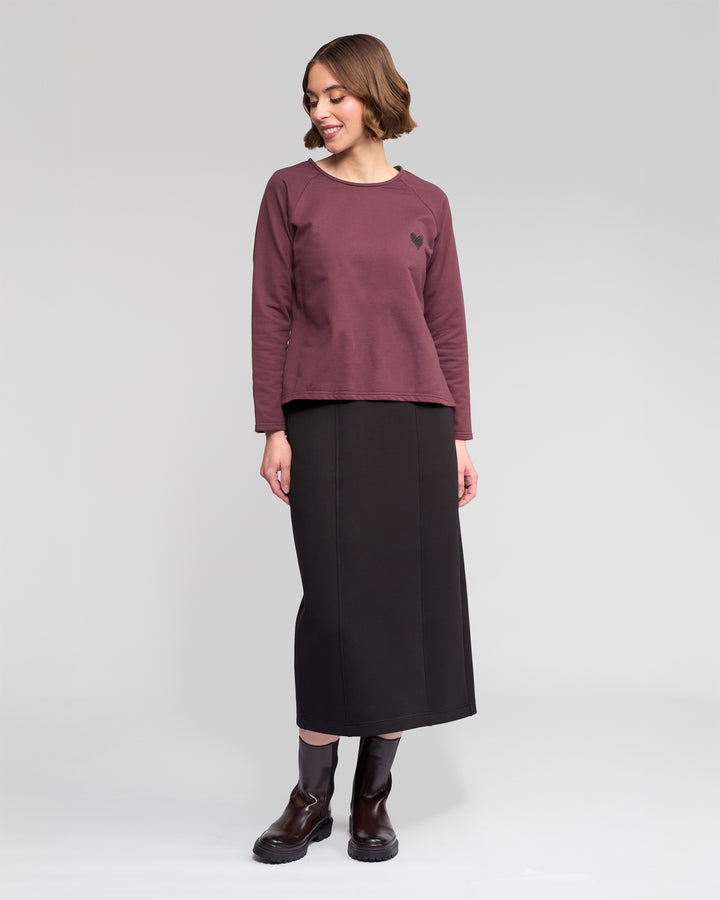 A woman stands in a studio wearing the Vassalli Lover Top - Port, a maroon relaxed long-sleeve with a small heart graphic, paired with a long black skirt and black boots. She looks down, smiling, her short brown hair styled in waves.