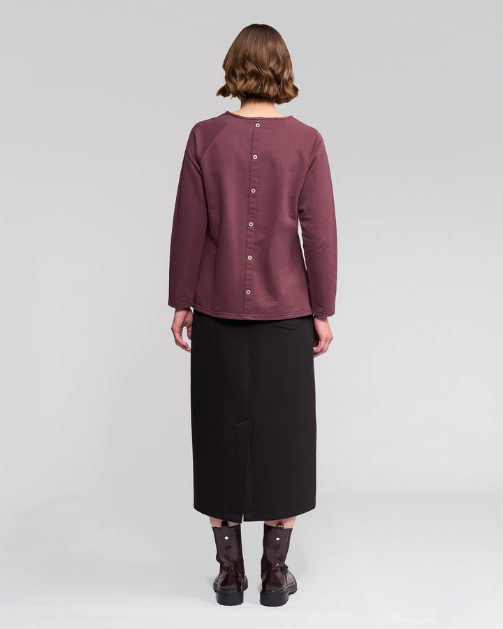 A person with short brown hair is seen from behind wearing the Vassalli Lover Top - Port, a relaxed long-sleeve top with back buttons, paired with a black midi skirt and dark ankle boots, standing against a light gray background.
