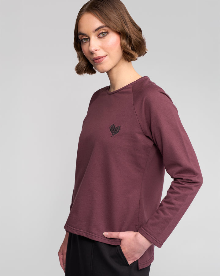A woman with short brown hair wears the Vassalli Lover Top in Port, a relaxed maroon cotton jersey with a small heart on the chest. She stands with one hand in her pocket, looking at the camera against a plain light gray background.