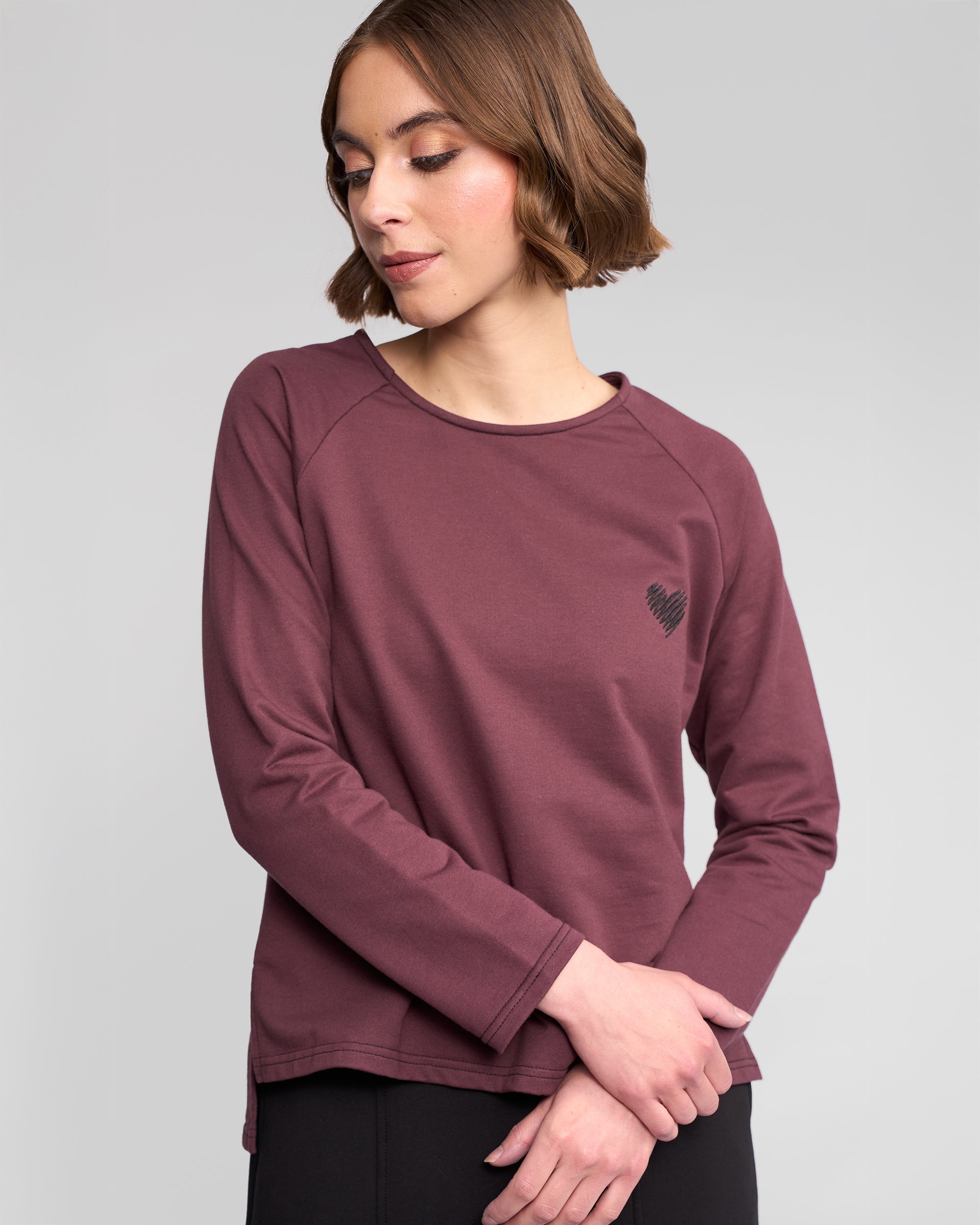 A woman with short brown hair wears the Vassalli Lover Top in Port, a relaxed maroon long-sleeve cotton jersey featuring a small black heart on the left chest, standing against a light gray background with her arms gently crossed.