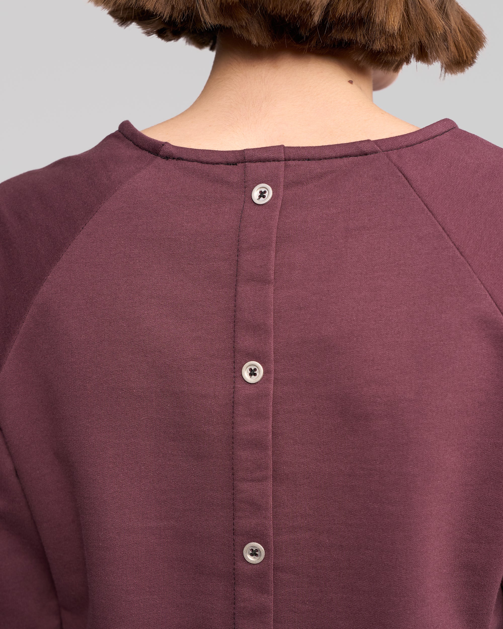 Close-up of a person with short brown hair wearing the Vassalli Lover Top - Port, a relaxed fit long-sleeve purple cotton jersey with visible seams and a row of buttons down the back.