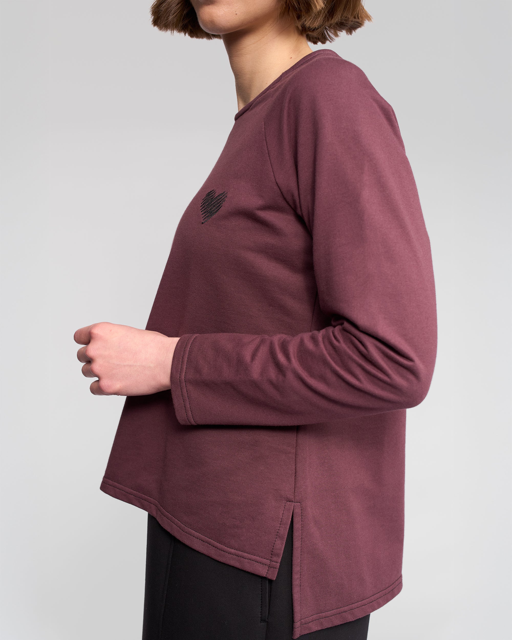 A person with short brown hair stands sideways against a plain light background, wearing the Vassalli Lover Top - Port, a relaxed burgundy long-sleeve featuring a small heart graphic on the chest.