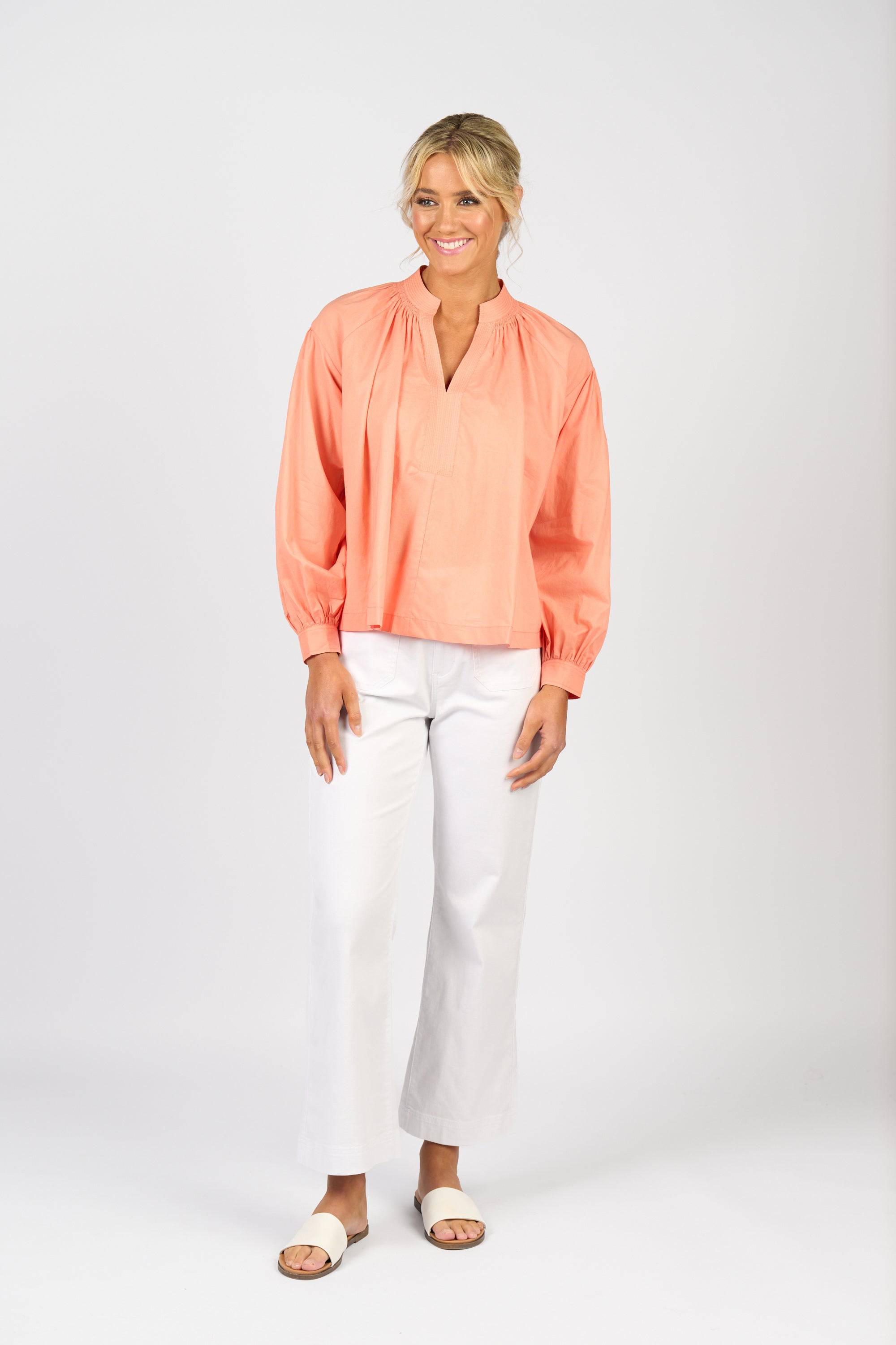 A woman smiles, wearing the Vassalli Placket Shirt in apricot with balloon sleeves and a v-neckline, paired with white pants and sandals against a plain white background.
