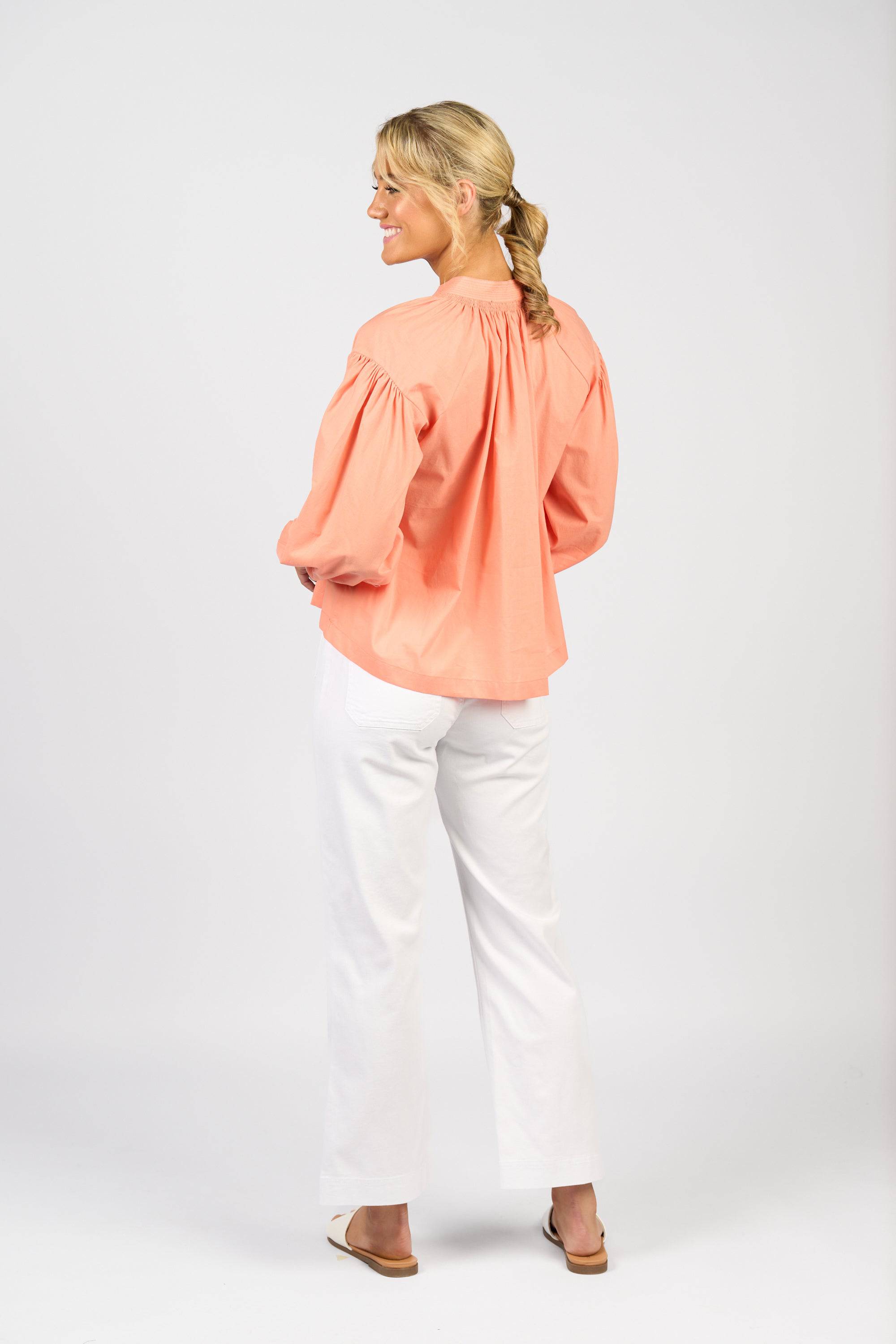 A woman with blond braided hair smiles and stands sideways, wearing the Vassalli Placket Shirt in Apricot with white pants and sandals against a plain light background.