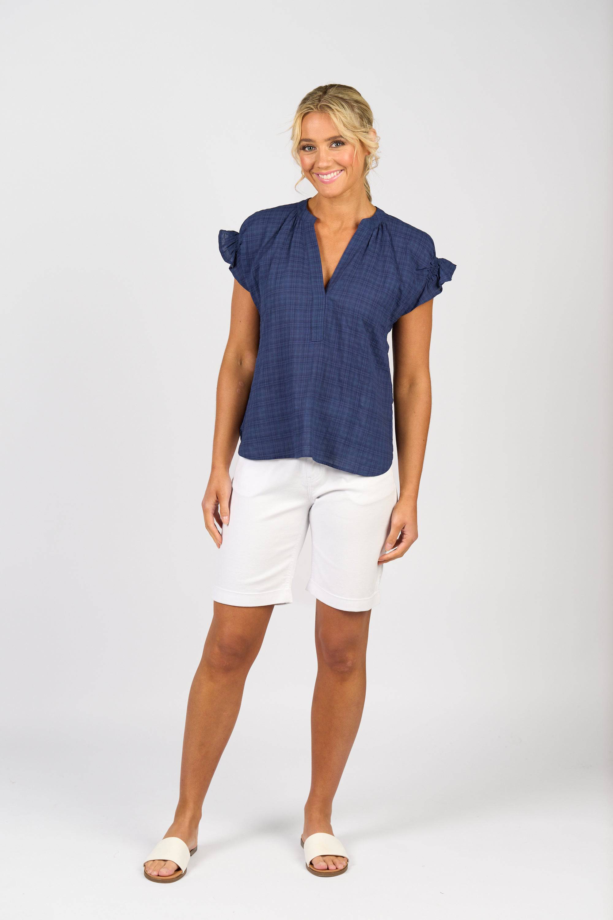A woman smiles while wearing the Vassalli Evelyn Top in navy, styled with white knee-length shorts and slide sandals, standing against a plain light background.