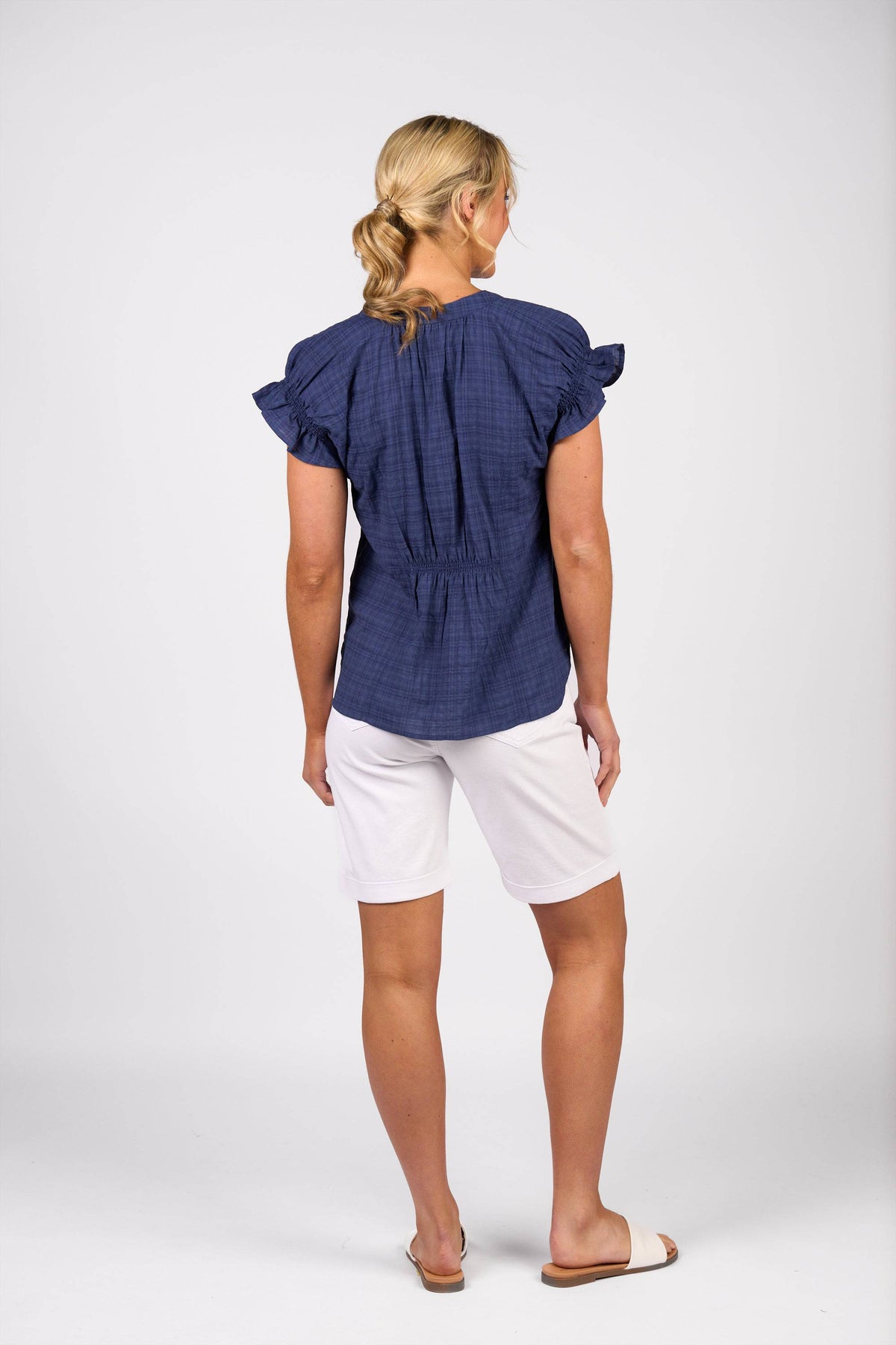 A woman with blonde hair tied back faces away, wearing the Vassalli Evelyn Top in navy with ruffled sleeves, white knee-length shorts, and white slide sandals against a plain light background.