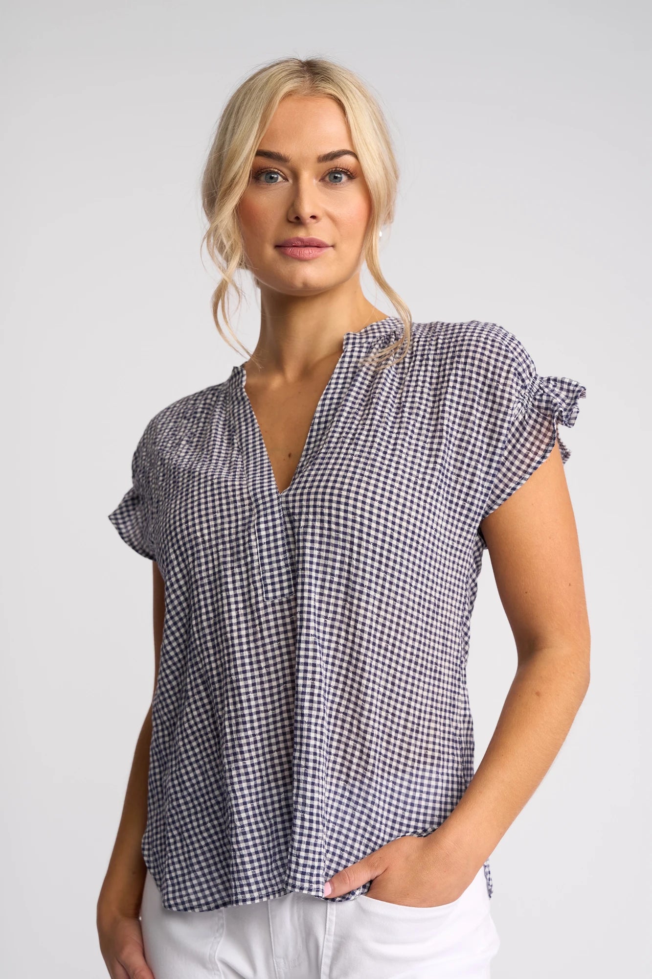 Wearing the Vassalli Evelyn Top in Ink Gingham—a relaxed-fit, blue and white cotton gingham design with a V-neckline—a blonde-haired woman stands against a plain light background, pairing the top with white pants and one hand in her pocket.
