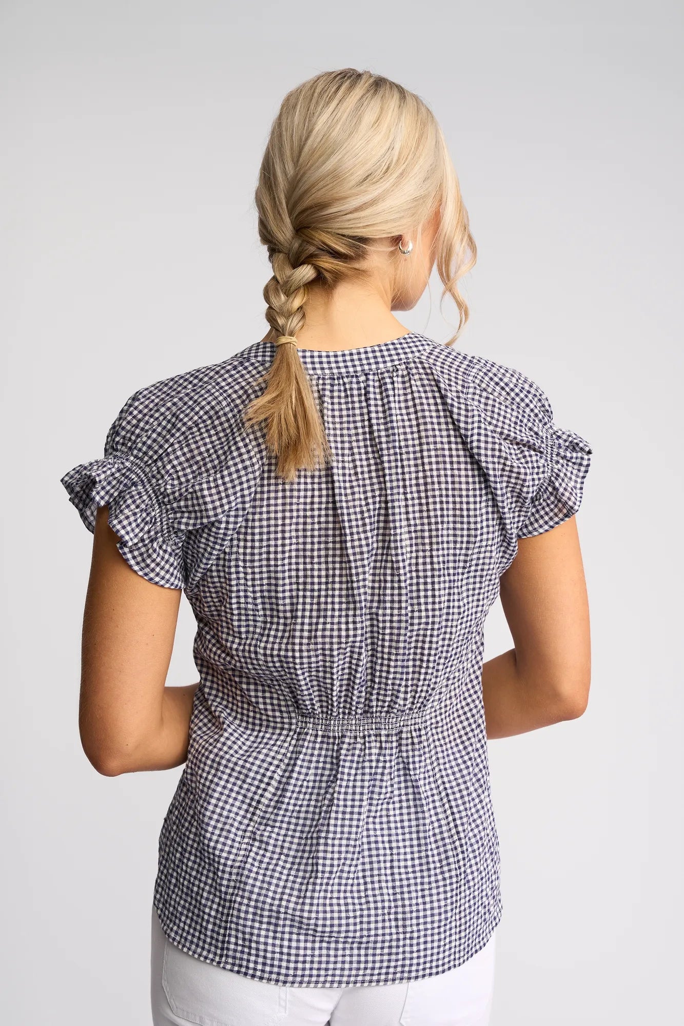 A woman with blonde hair in a braid is shown from behind, wearing the Vassalli Evelyn Top in Ink Gingham—a short-sleeve, blue and white cotton blouse with ruffled sleeves—paired with white pants. The relaxed silhouette of the top lends a breezy charm to her look.