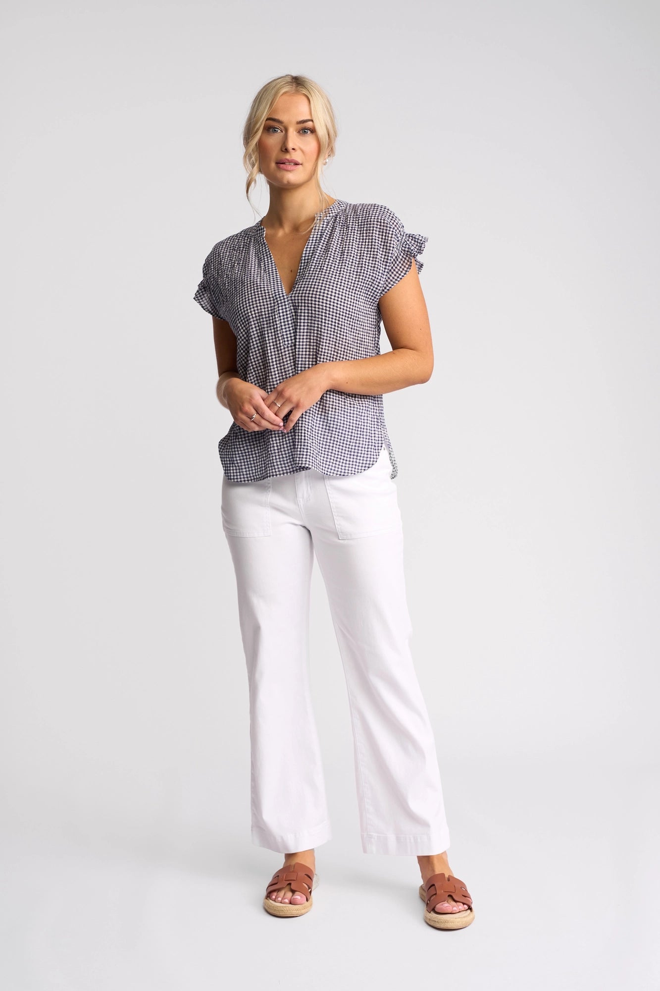 A woman stands against a plain light background, wearing the Vassalli Evelyn Top in Ink Gingham—a short-sleeve cotton top with a relaxed silhouette—paired with white pants and brown sandals. She has blonde hair styled in a loose updo and a neutral expression.