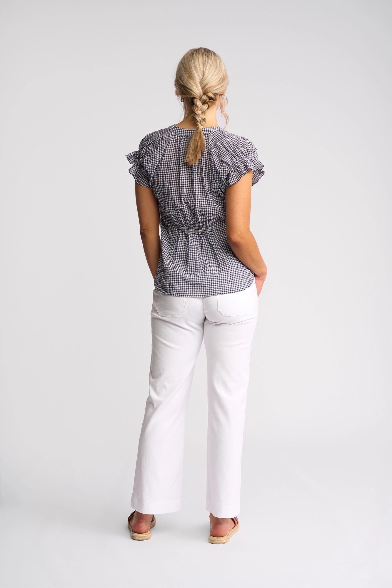A woman with blonde hair in a braid stands facing away, wearing the Vassalli Evelyn Top in Ink Gingham—a relaxed silhouette cotton top with short sleeves—paired with white pants and sandals against a plain white background.
