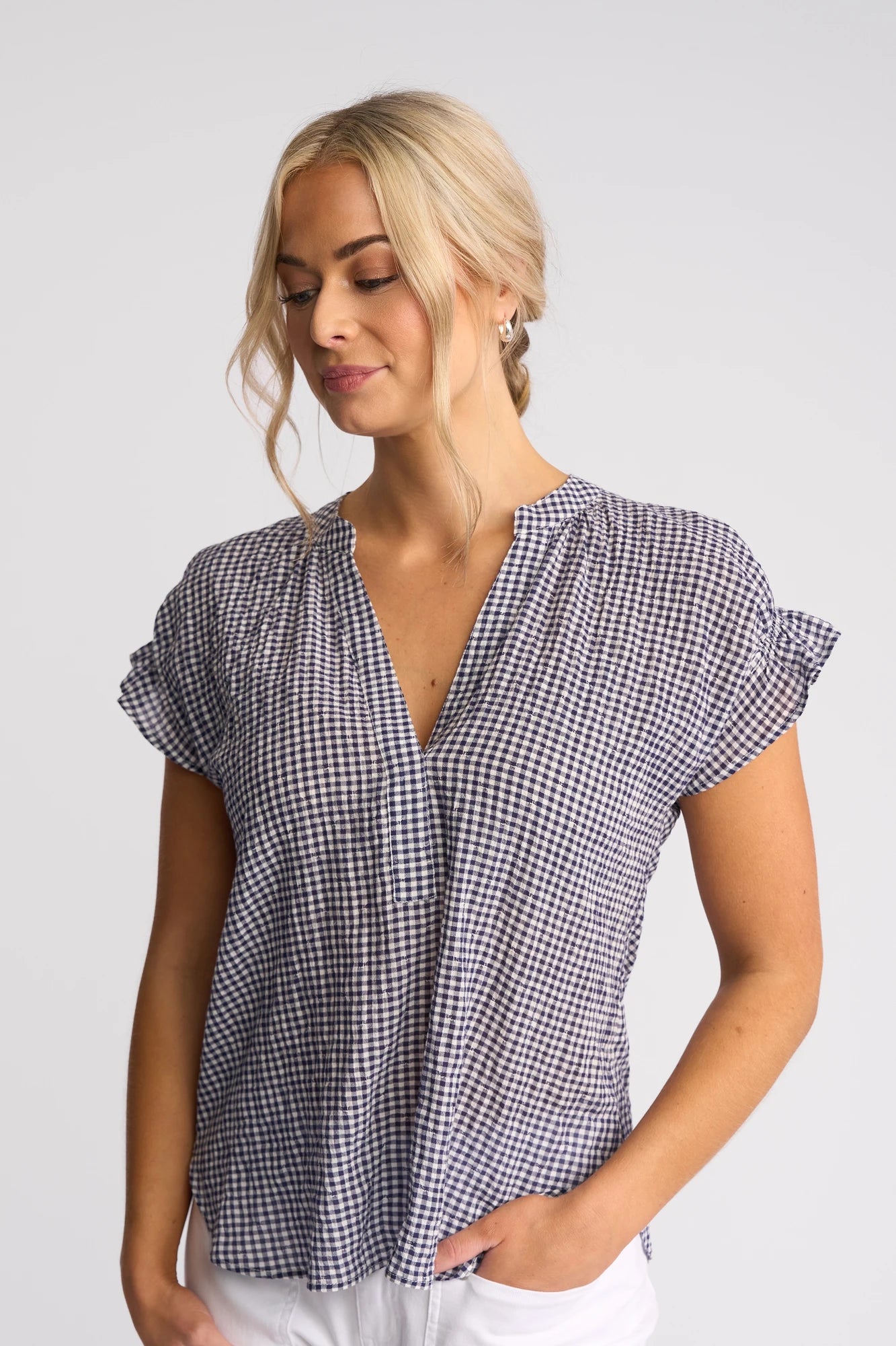 A woman with blonde hair in a loose braid wears the Vassalli Evelyn Top – Ink Gingham, a short-sleeved black-and-white cotton gingham top featuring a V-neckline and relaxed silhouette, paired with white pants. She stands against a plain light background with one hand in her pocket, looking slightly down and smiling.