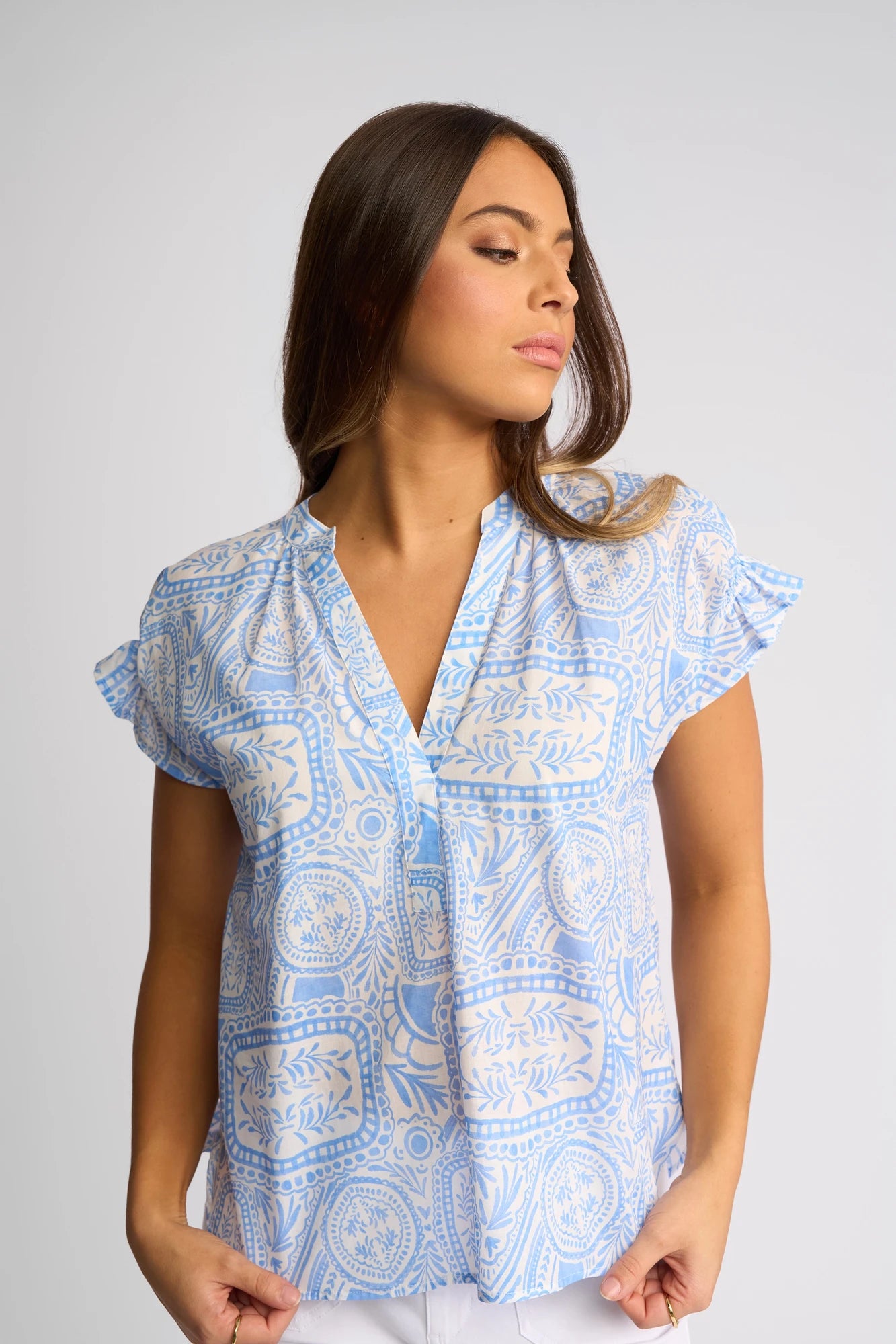 Woman with long brown hair wearing a lightweight cotton Vassalli Evelyn Top - Palma in a light blue and white Mediterranean tile print, looking to her left, standing against a plain light background.