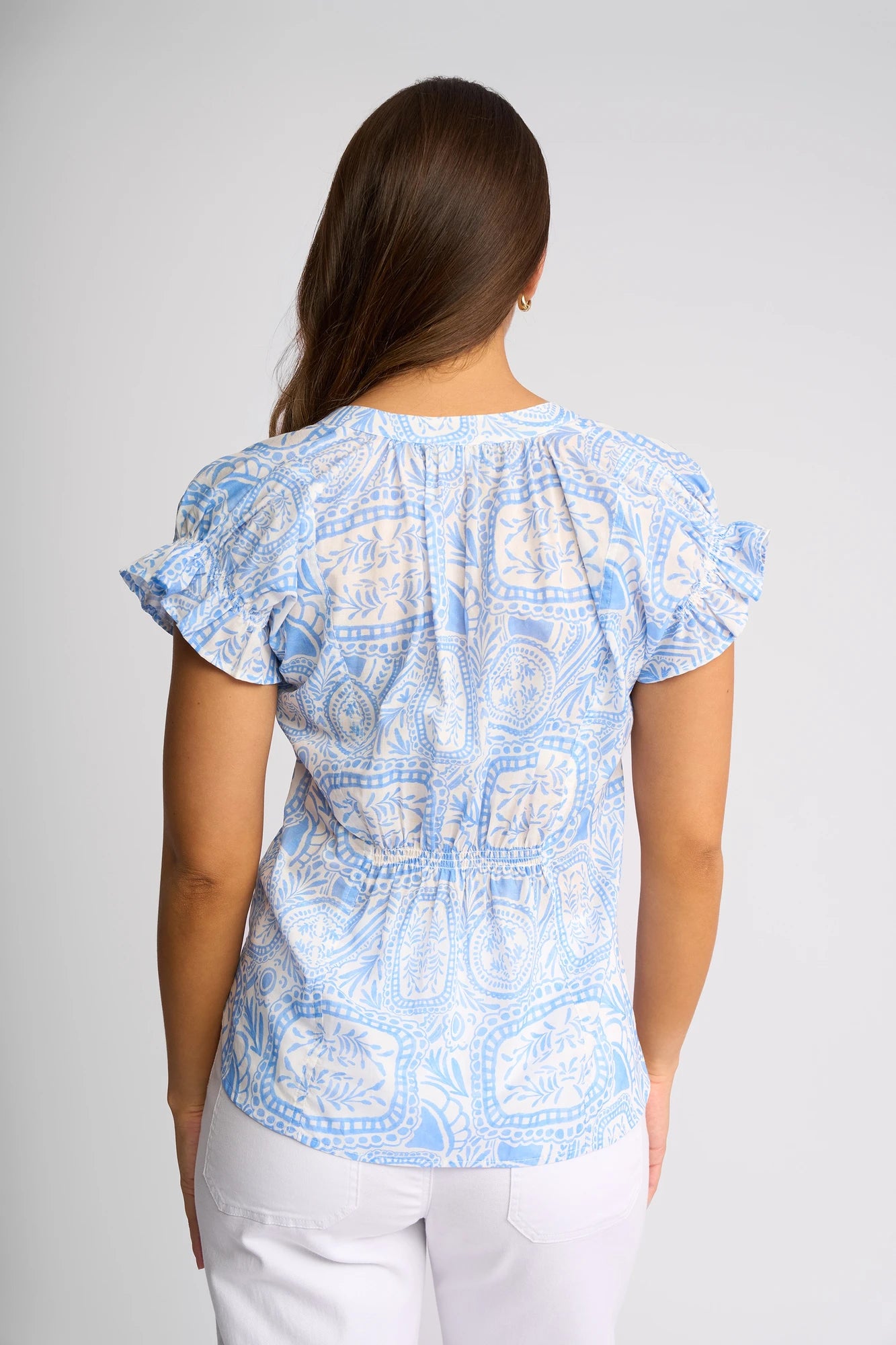 A woman with long brown hair is seen from behind, dressed in the Vassalli Evelyn Top - Palma, a light blue mediterranean tile print blouse with flutter sleeves made from lightweight cotton, paired with white pants against a plain background.