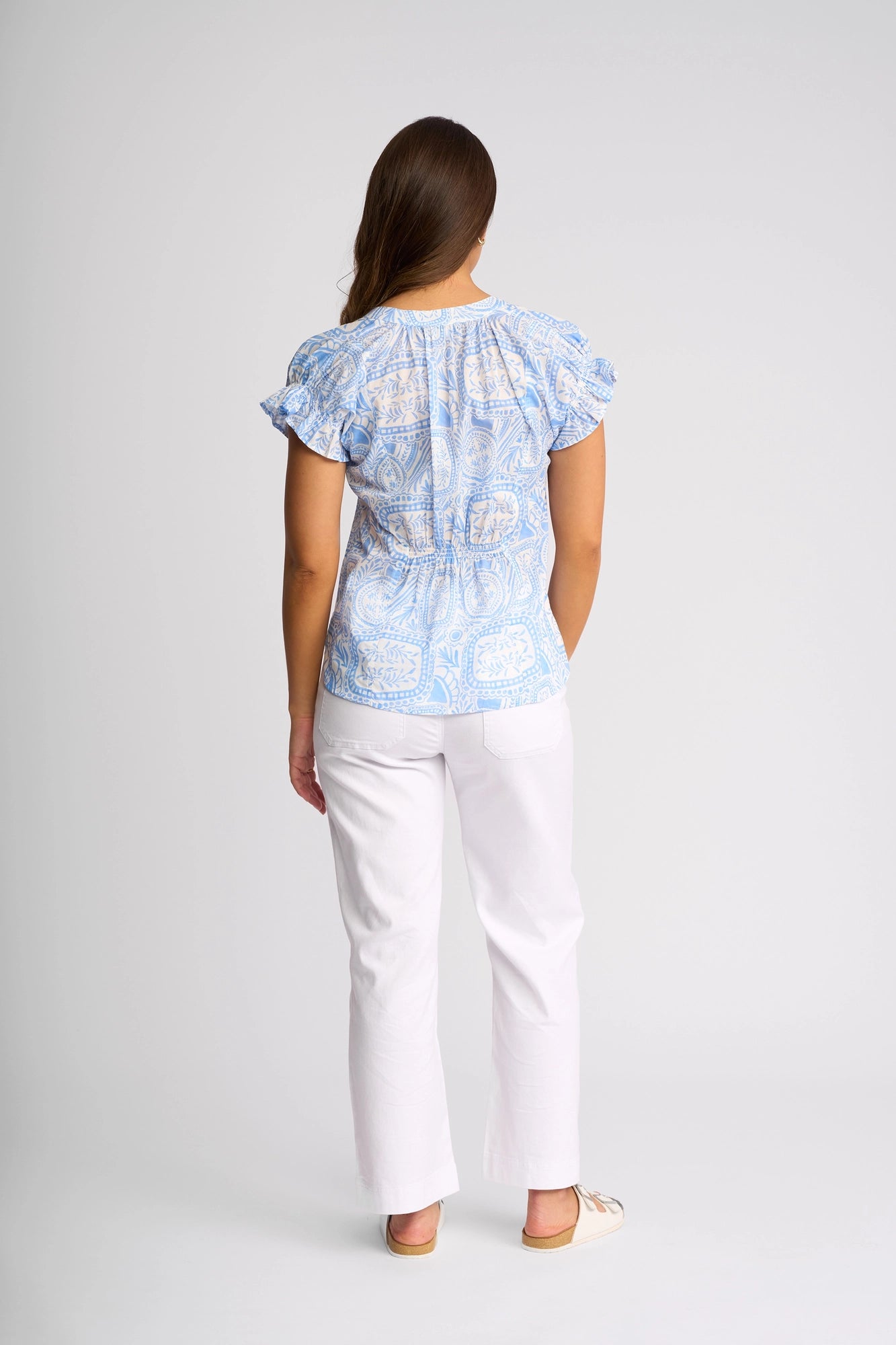 A woman stands facing away, wearing the Vassalli Evelyn Top - Palma—a short-sleeved blouse with a blue Mediterranean tile-inspired print—paired with white pants and beige flats, set against a plain light background.