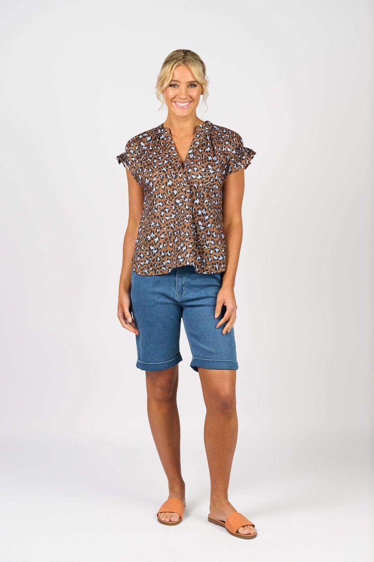 A woman stands smiling against a plain white background, wearing the Vassalli Evelyn Top - Nala with a brown and black leopard print, blue knee-length denim shorts, and tan sandals.