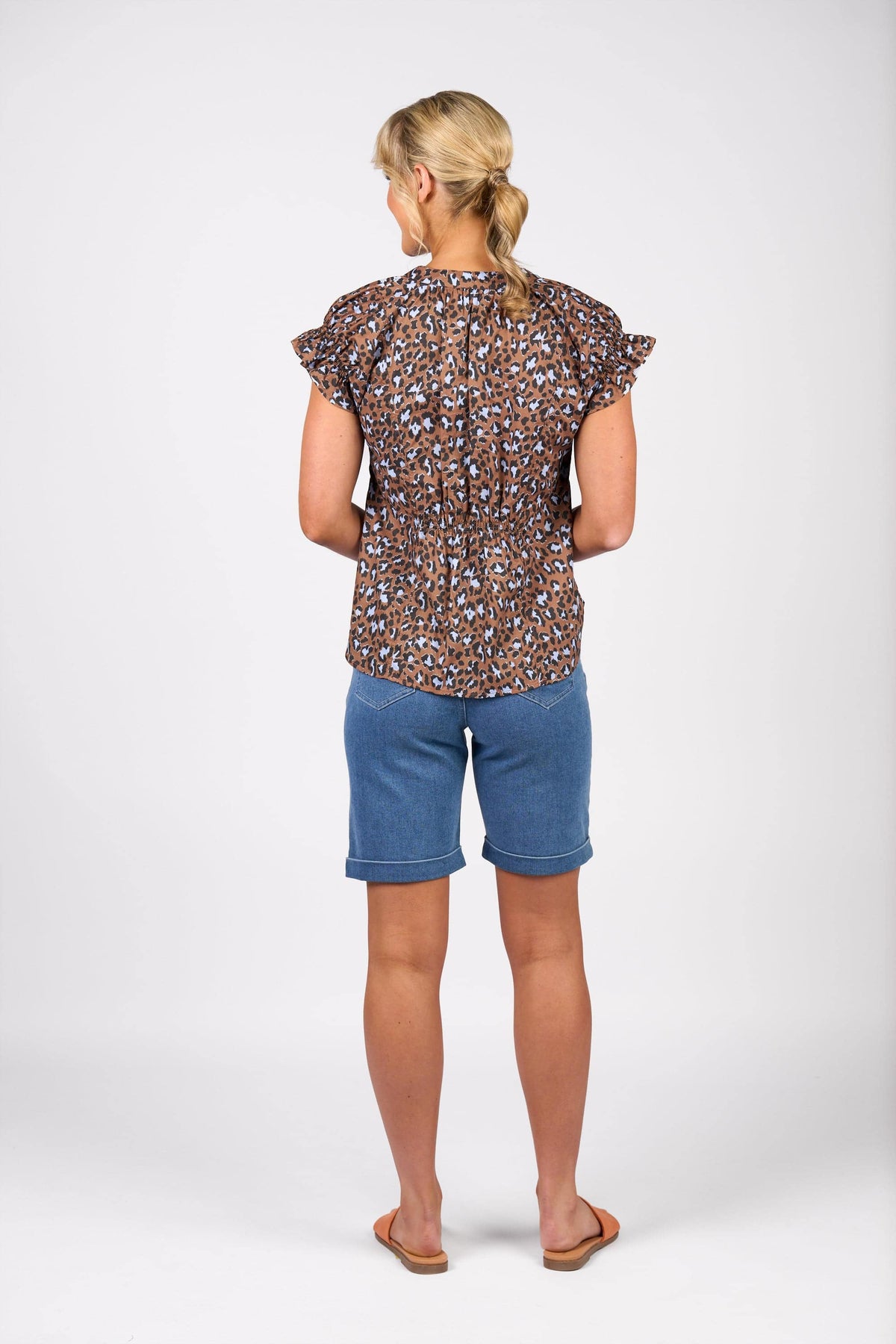 A woman with blonde hair in a low ponytail stands facing away, wearing the Vassalli Evelyn Top - Nala, featuring a brown and blue leopard print with ruffled sleeves, paired with blue denim shorts and tan sandals.