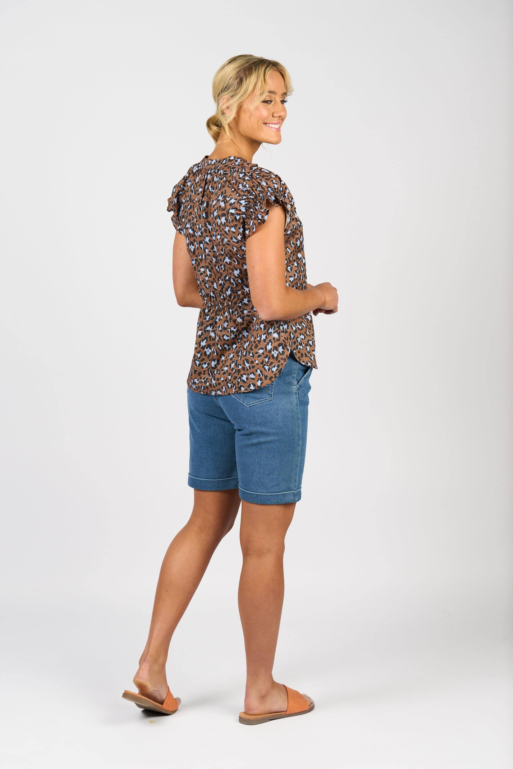 A woman smiles, turned slightly to the side, wearing the Vassalli Evelyn Top - Nala with a brown pattern, paired with blue denim shorts and brown sandals. Her blonde hair is tied back against a plain white background.
