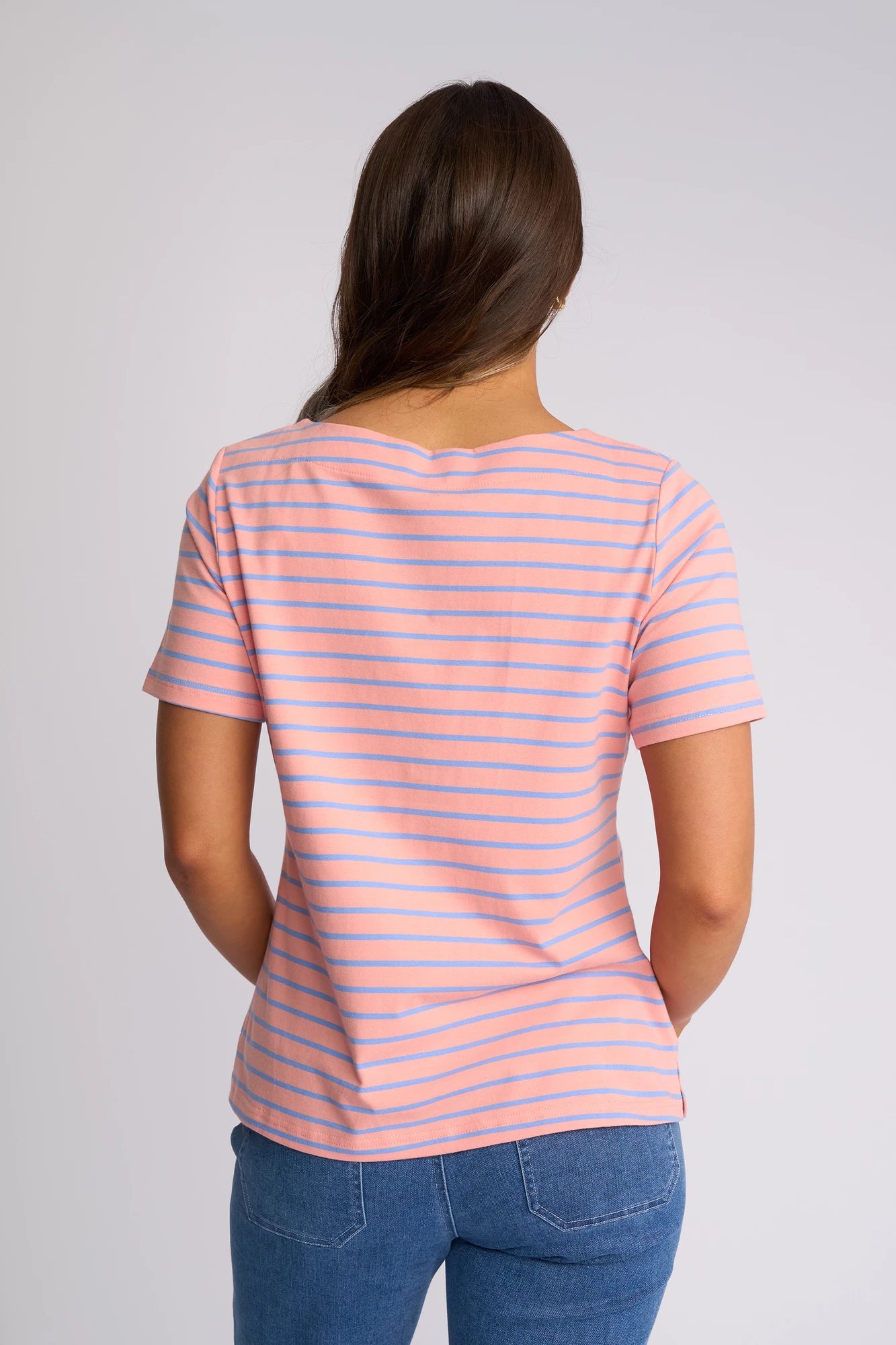 A person with long brown hair is standing with their back to the camera, wearing the Vassalli Raya Top in Apricot/Blue, which features a pink and blue striped design and a flattering boat neckline. They have paired it with blue jeans against a plain light background.