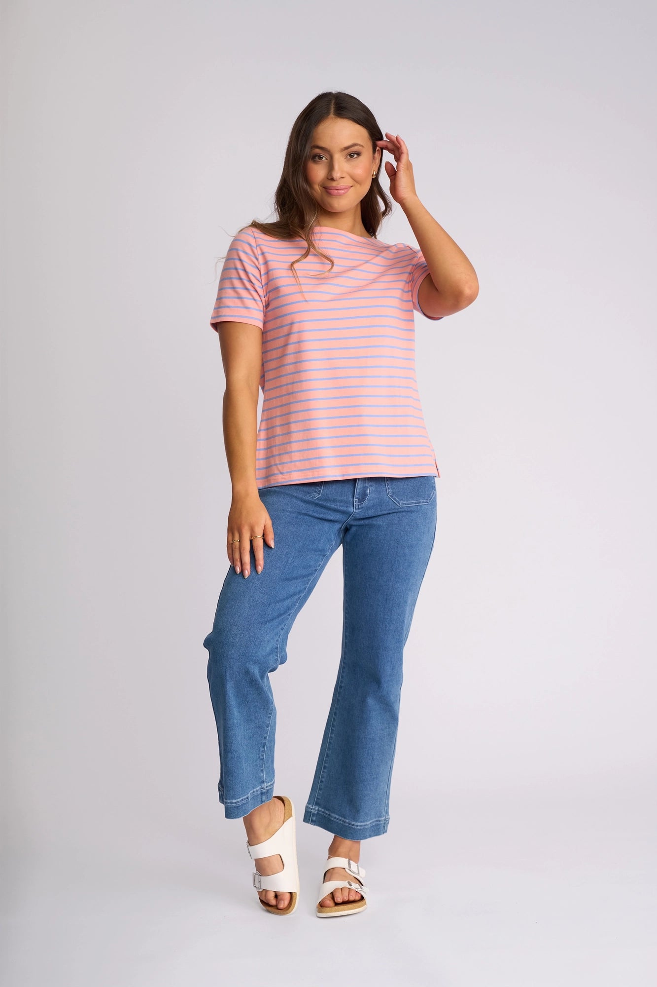 A woman stands against a plain light background, smiling and touching her hair with one hand, wearing the Vassalli Raya Top in Apricot/Blue—a stretch cotton piece featuring pink and orange stripes and a boat neckline—paired with blue jeans and white sandals. This top combines comfort with effortless style.