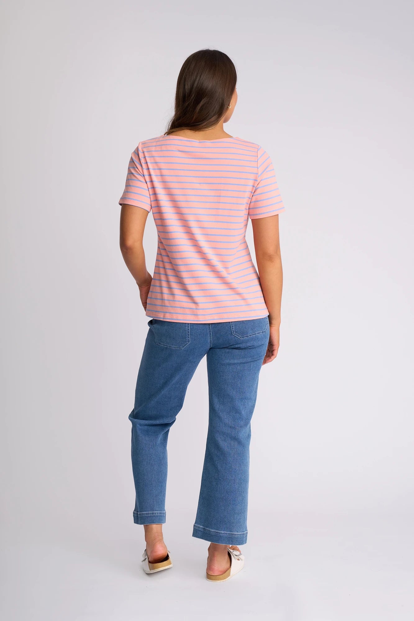 A woman stands facing away, wearing a Vassalli Raya Top in Apricot/Blue with a boat neckline, paired with blue jeans and white shoes against a plain white background.