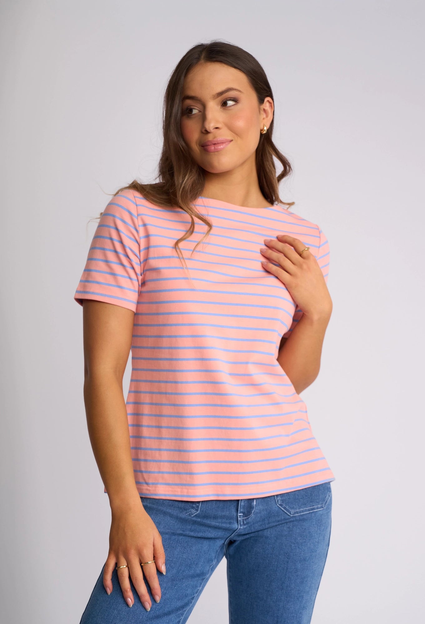 A woman wearing a Vassalli Raya Top in Apricot/Blue—a stretch cotton top featuring pink and blue stripes and a flattering boat neckline—pairs it with blue jeans as she stands against a light background, gazing to the side with a slight smile and gently touching her shirt with one hand.