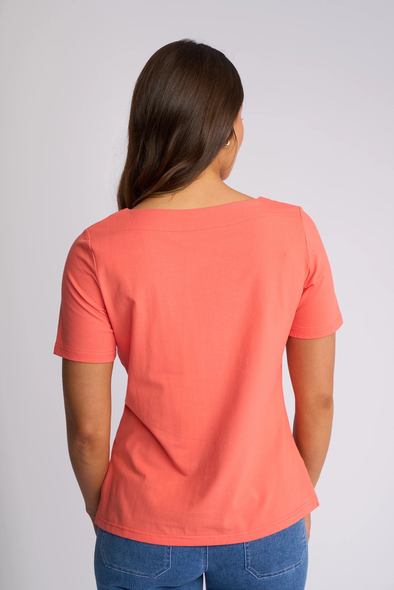 A woman with long brown hair, seen from behind, is wearing the Vassalli Raya Top in Coral, featuring a stylish boat neckline, paired with blue jeans against a plain light background.