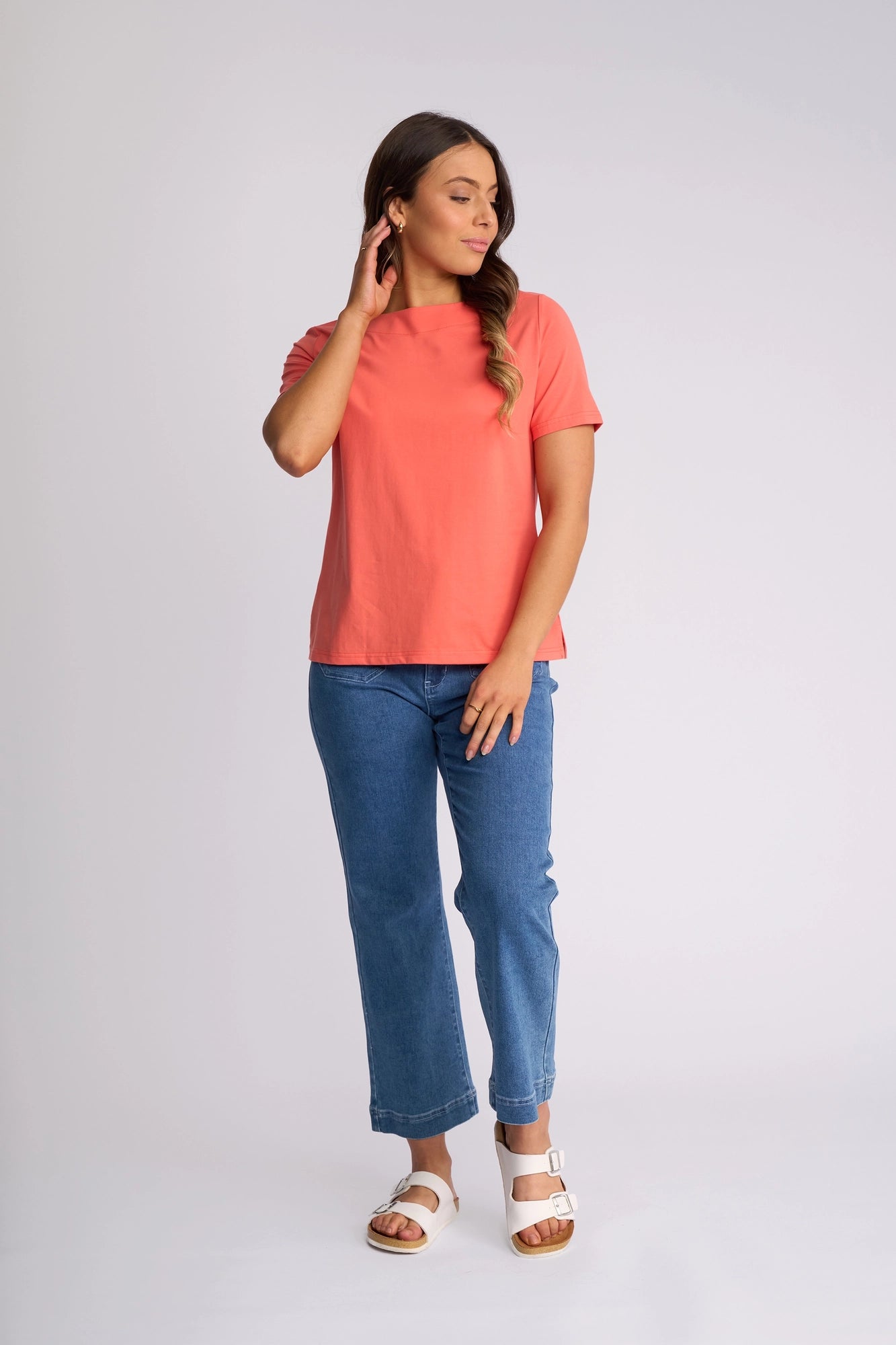 A woman stands against a plain background, wearing the Vassalli Raya Top in Coral, which features a stylish boat neckline. She pairs it with blue jeans and white sandals, posing with one hand touching her hair and the other resting on her thigh.