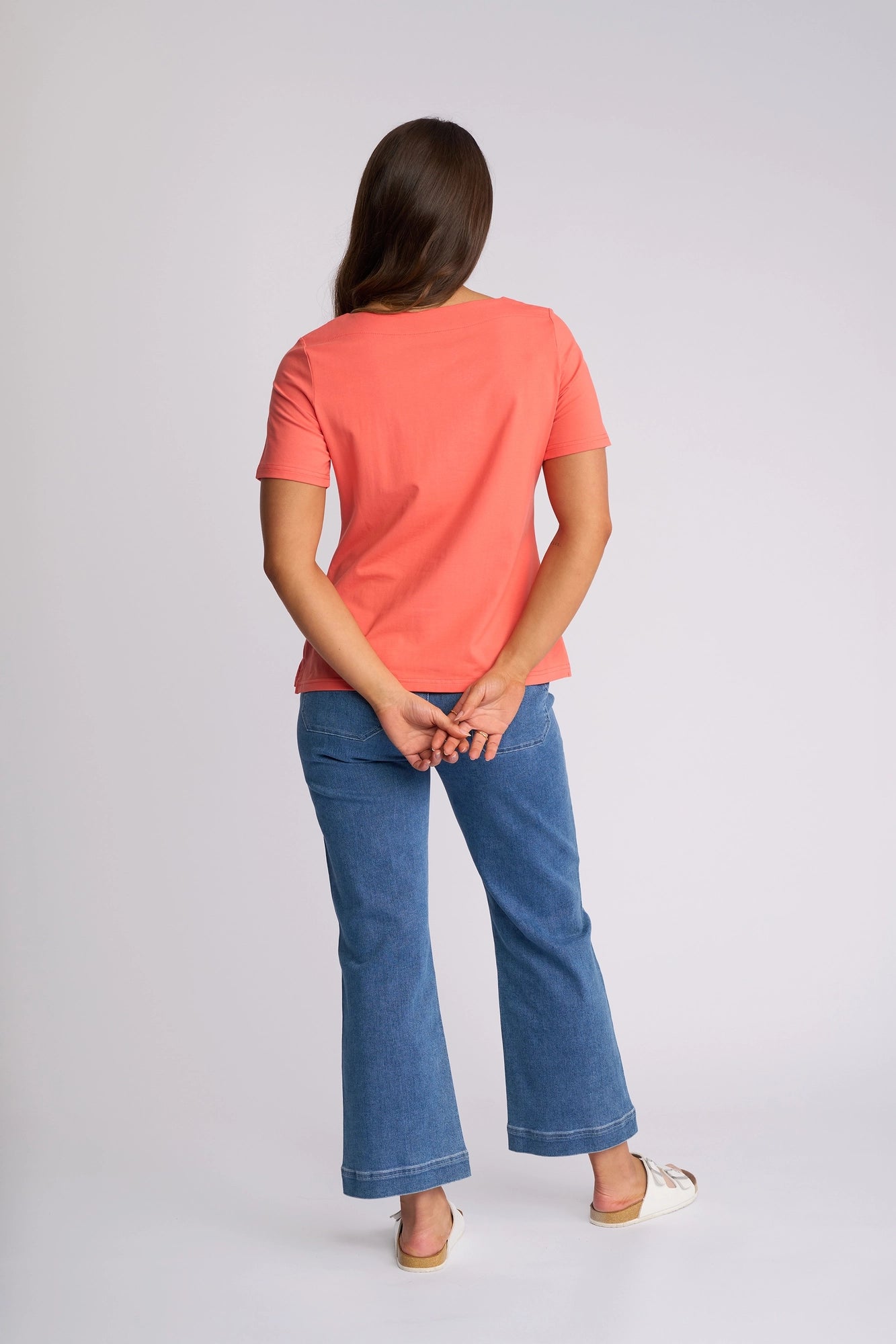 A woman stands facing away with her hands clasped behind her back. She wears the Vassalli Raya Top in coral, featuring a stretch cotton fabric and boat neckline, paired with blue jeans and white shoes, against a plain light background.