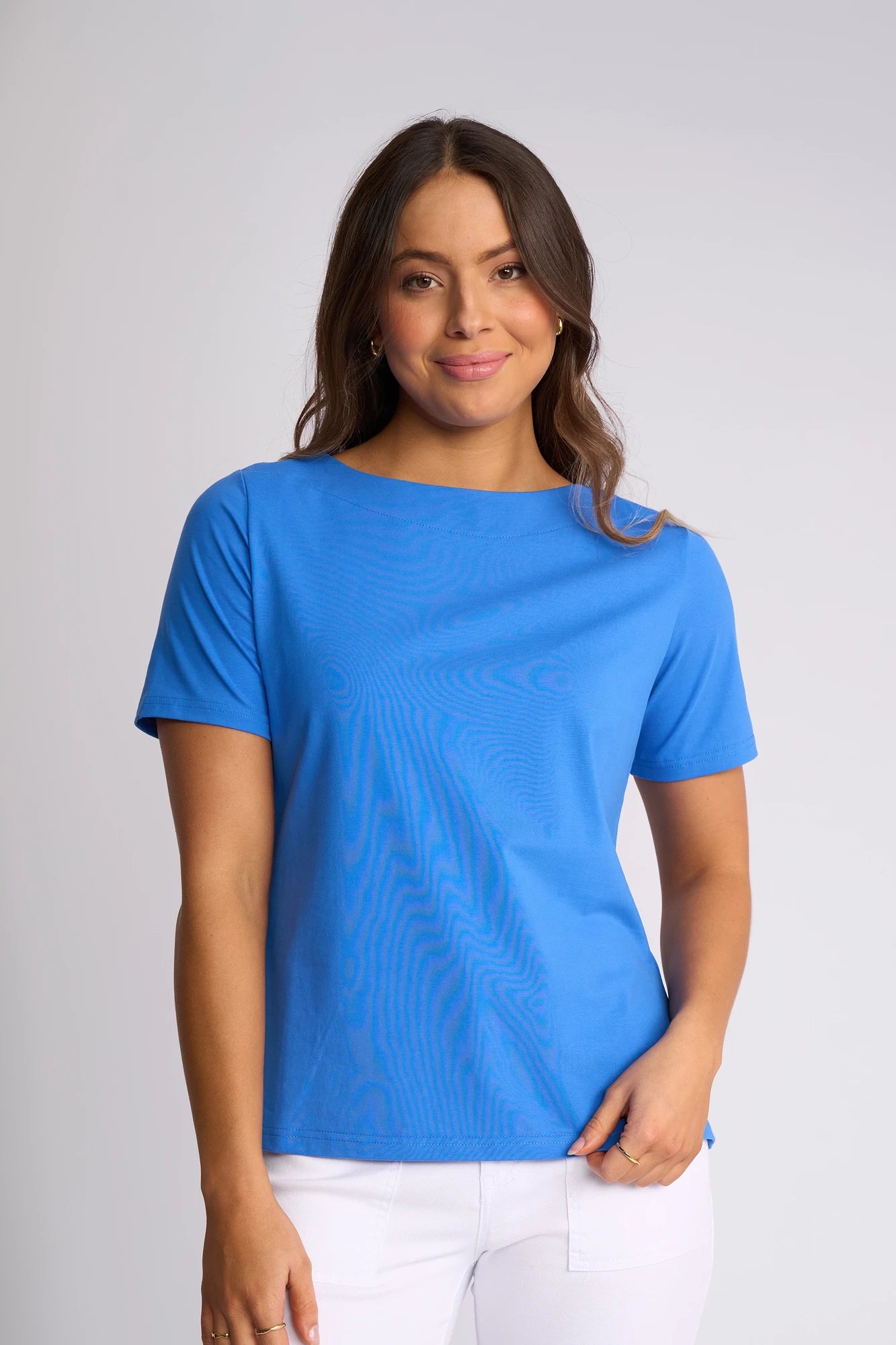 A woman with long brown hair wears the Vassalli Raya Top in French Blue, featuring stretch cotton fabric and a stylish boat neckline. She pairs it with white pants and stands against a light gray background, smiling softly at the camera.