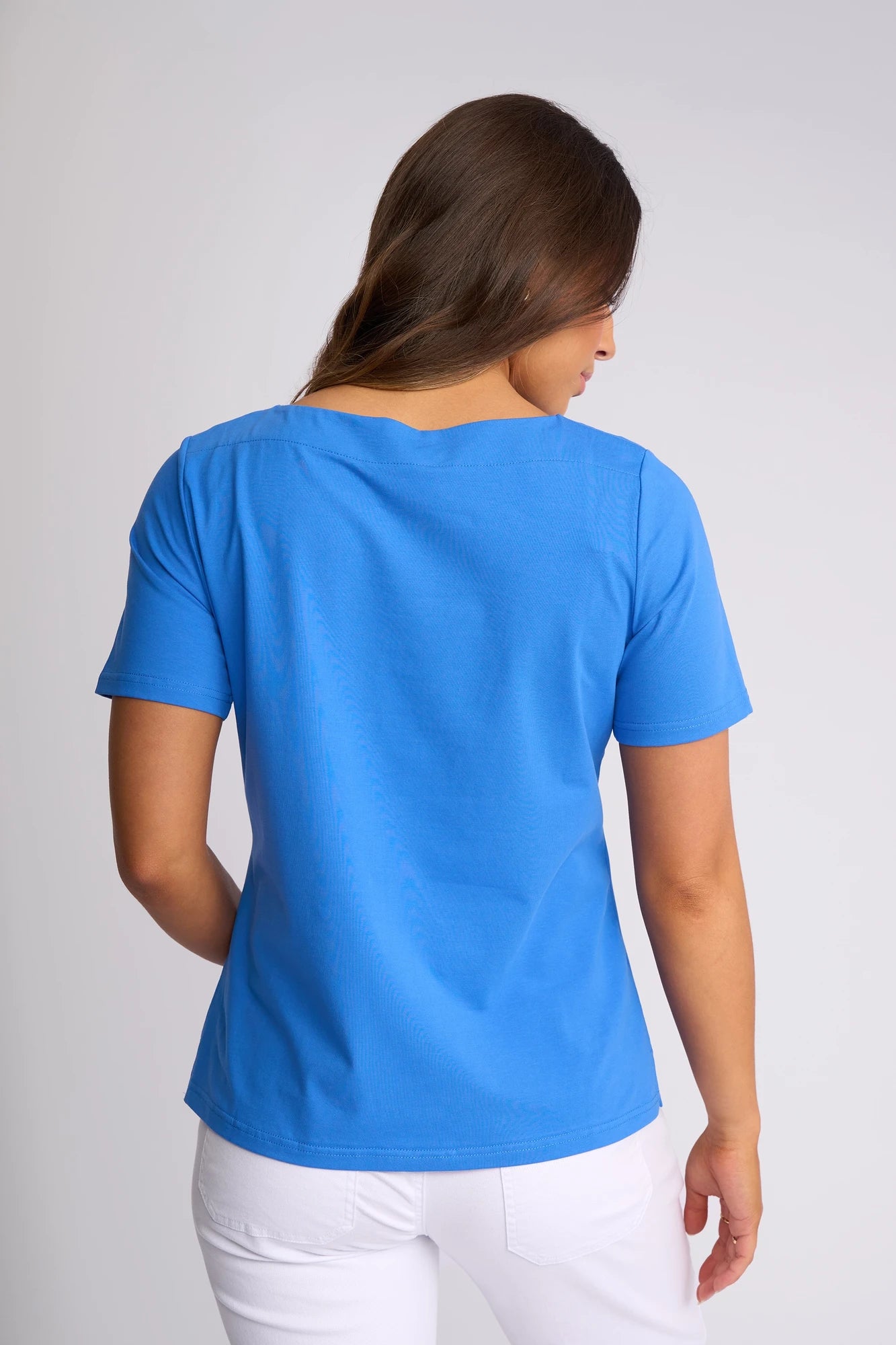 A woman with long brown hair, wearing the Vassalli Raya Top in French Blue featuring a boat neckline and paired with white pants, stands facing away from the camera against a light gray background.
