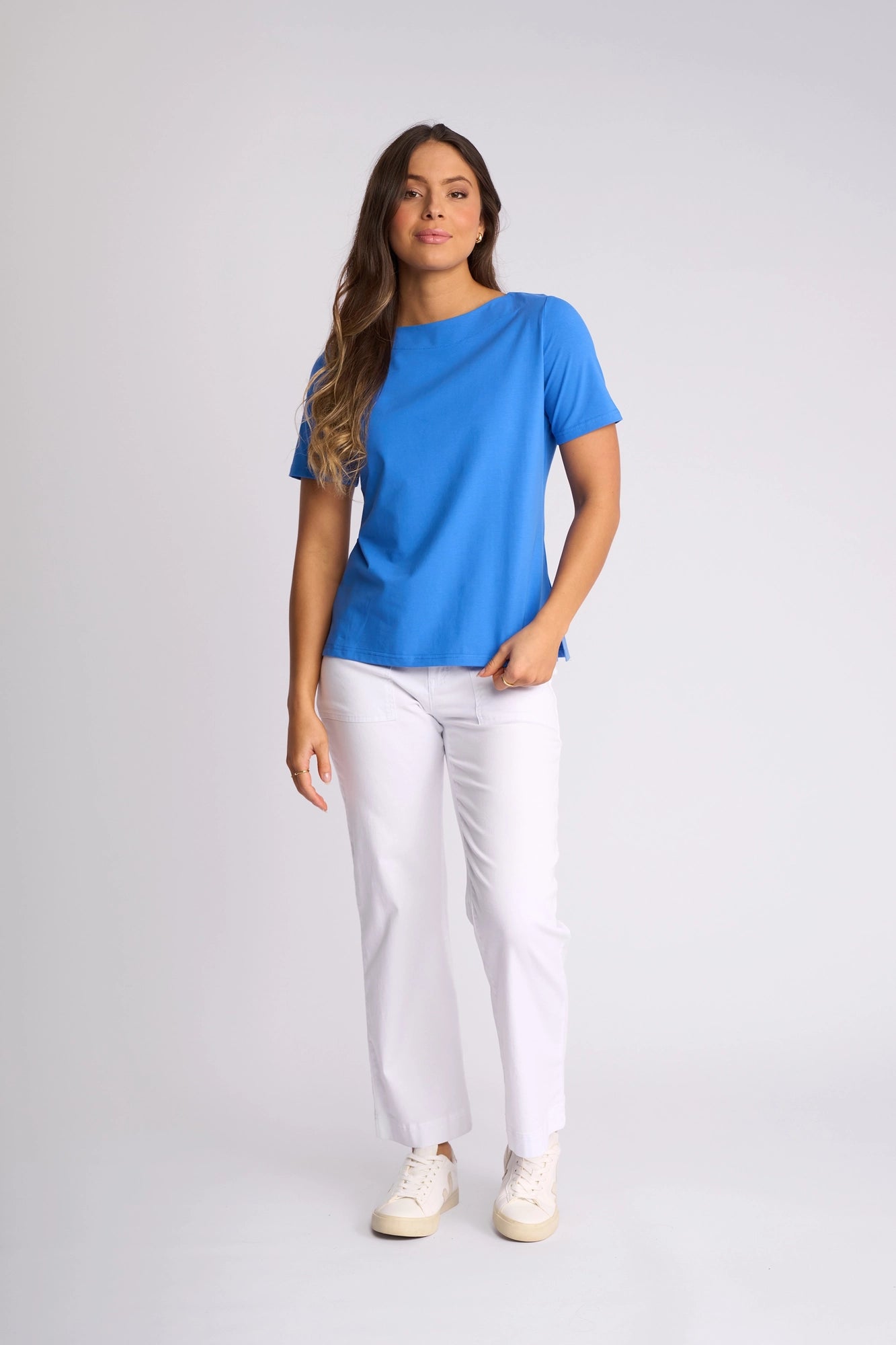 A woman poses in a studio wearing the Vassalli Raya Top in French Blue—a vibrant, stretch cotton short-sleeve top featuring a boat neckline—styled with white pants and white sneakers. She has long, wavy brown hair and stands against a plain light gray background.