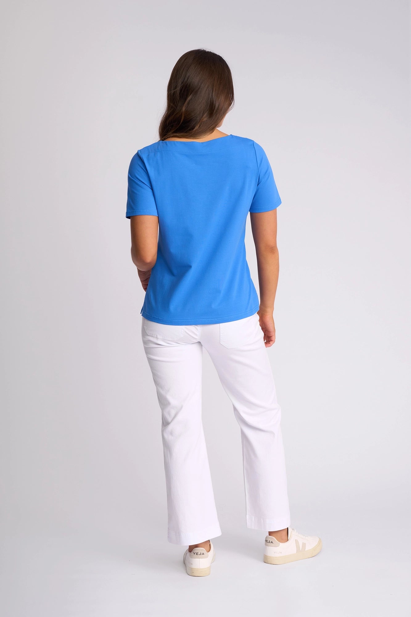 A person with long brown hair stands facing away, wearing the Vassalli Raya Top in French Blue—crafted from stretch cotton with a stylish boat neckline—paired with white pants and white sneakers against a plain light background.