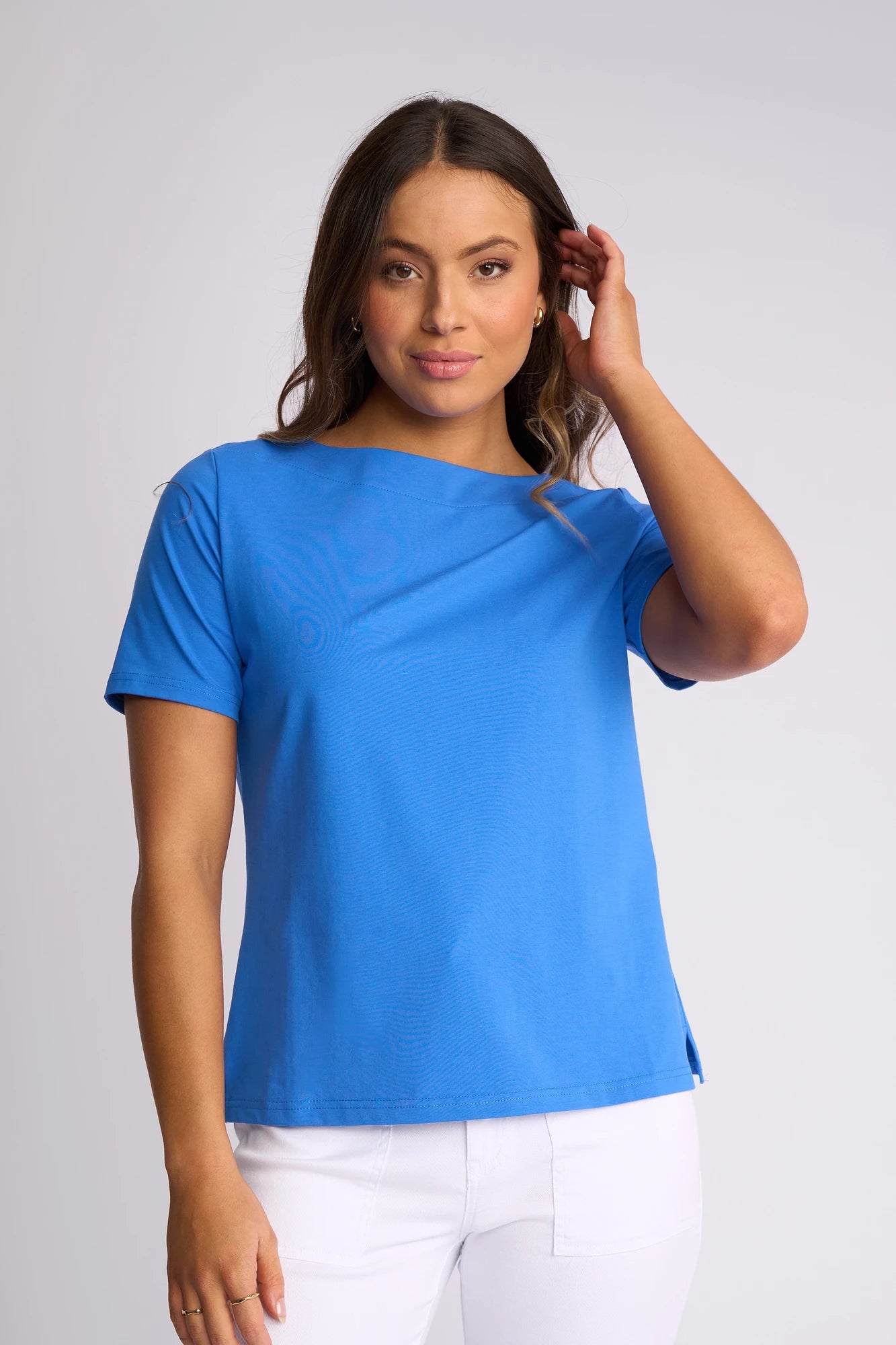 A woman stands against a plain light gray background, touching her hair and looking at the camera while wearing the Vassalli Raya Top in French Blue, featuring a boat neckline and stretch cotton fabric.