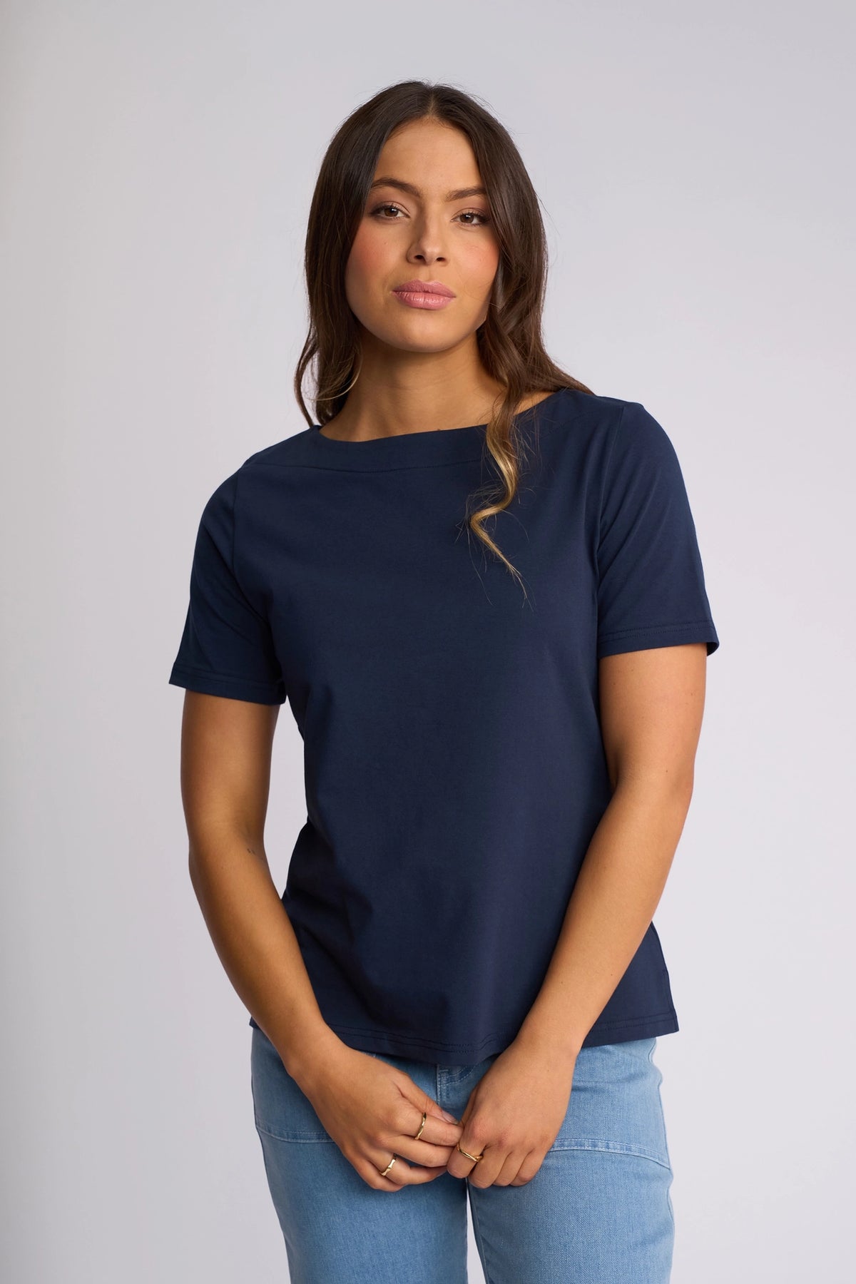 A woman with long brown hair, wearing the Vassalli Raya Top in Ink and light blue jeans, stands against a plain light background, looking at the camera with a neutral expression.
