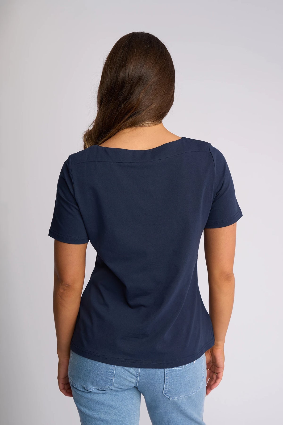 A woman with long brown hair, wearing the Vassalli Raya Top - Ink, which features a boat neckline and is crafted from stretch cotton for comfort and style, pairs it with light blue jeans as she stands facing away from the camera against a plain background.