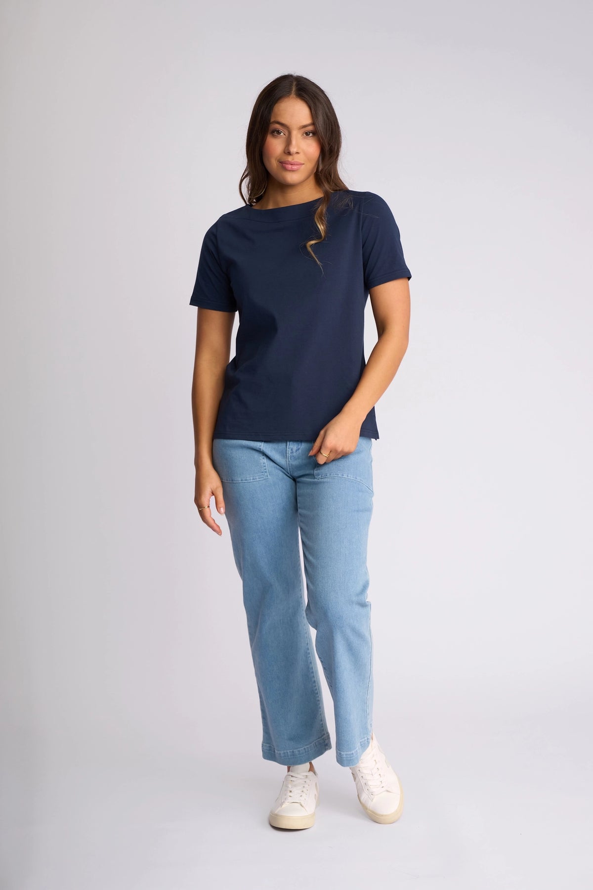 A person stands against a plain background, wearing the Vassalli Raya Top in Ink with a boat neckline, paired with light blue jeans and white shoes. They have long, wavy hair and a relaxed posture, with one hand resting on their thigh.