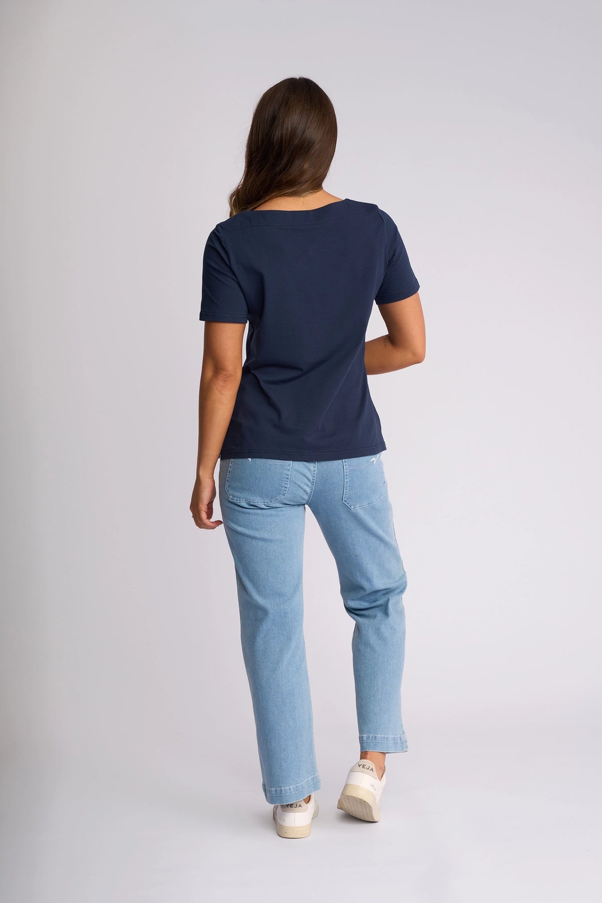 A woman with long brown hair, wearing the Vassalli Raya Top in Ink—a stretch cotton top with a boat neckline—paired with light blue jeans and white shoes, stands facing away from the camera against a plain light background.