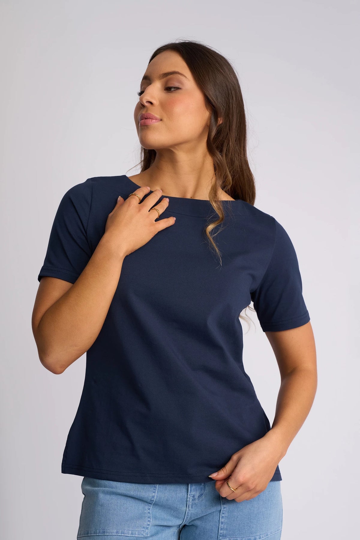 A woman with long brown hair wears the Vassalli Raya Top - Ink, featuring a stretch cotton fabric and boat neckline, paired with light blue jeans. She stands against a plain light background, with one hand near her neck and the other at her hip.