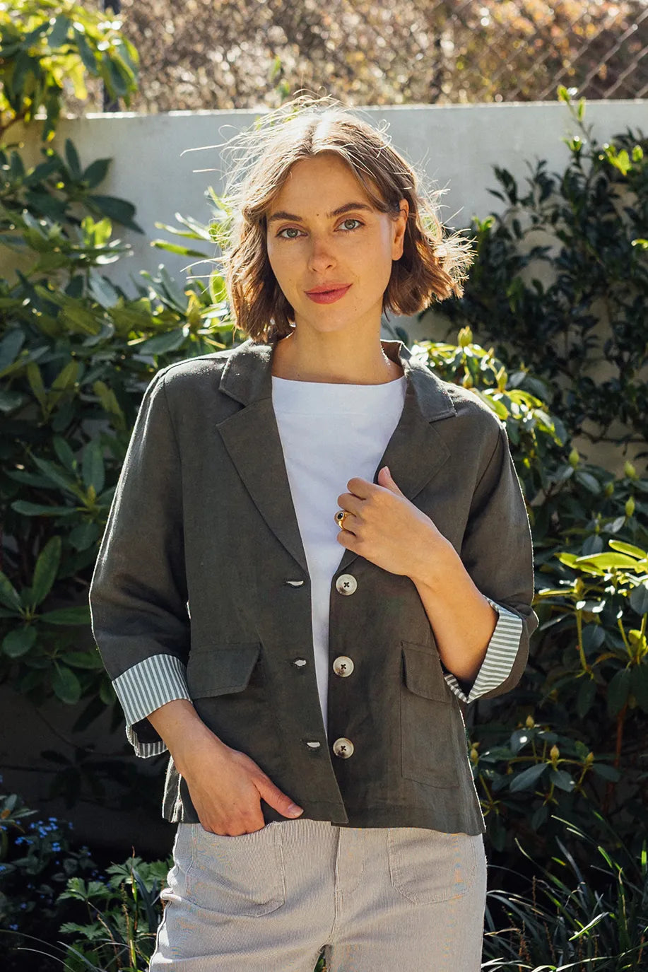 A woman with short brown hair stands outdoors, wearing the Vassalli Honey Jacket in khaki over a white shirt and light pants. She poses with one hand lightly touching her blazer, surrounded by green plants and sunlight.