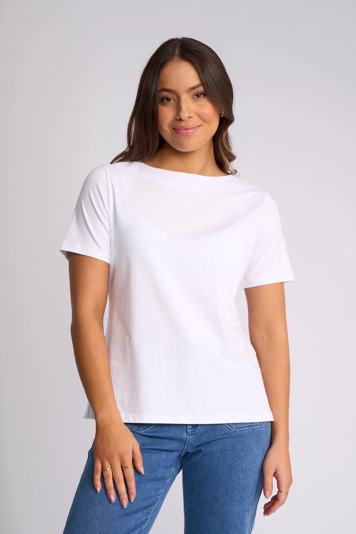 A woman with long brown hair wears the Vassalli Raya Top in white—a stretch cotton short-sleeve t-shirt—paired with blue jeans, standing against a light gray background and smiling slightly at the camera.