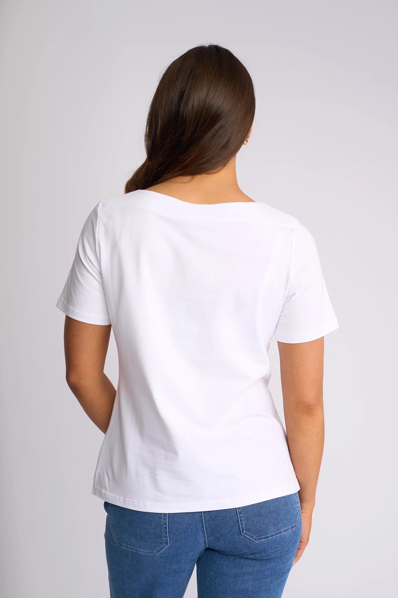 A person with long brown hair stands facing away, wearing the Vassalli Raya Top in White, crafted from stretch cotton, paired with blue jeans against a light gray background.