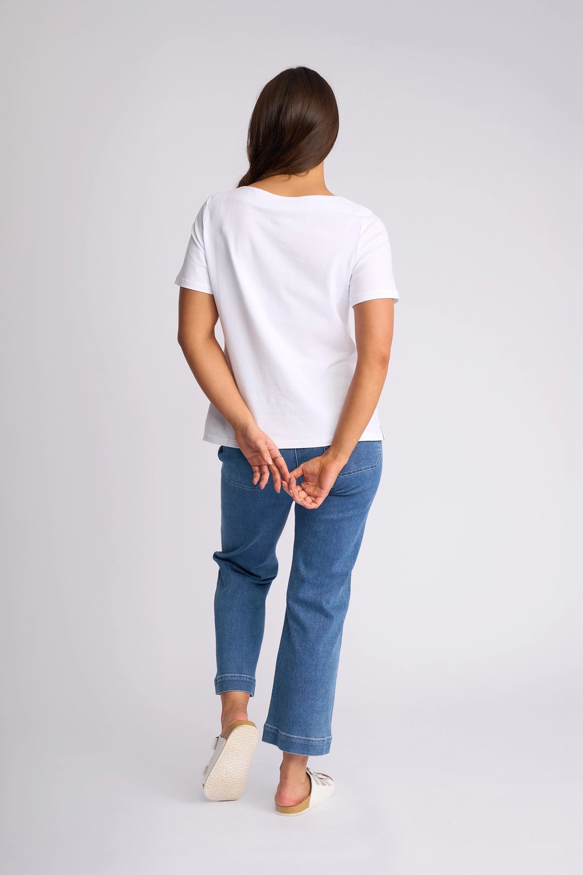 A person stands facing away against a plain light background, wearing the Vassalli Raya Top in white—a stretch cotton t-shirt with a boat neckline—paired with blue jeans and white slippers, their hands clasped behind their back.